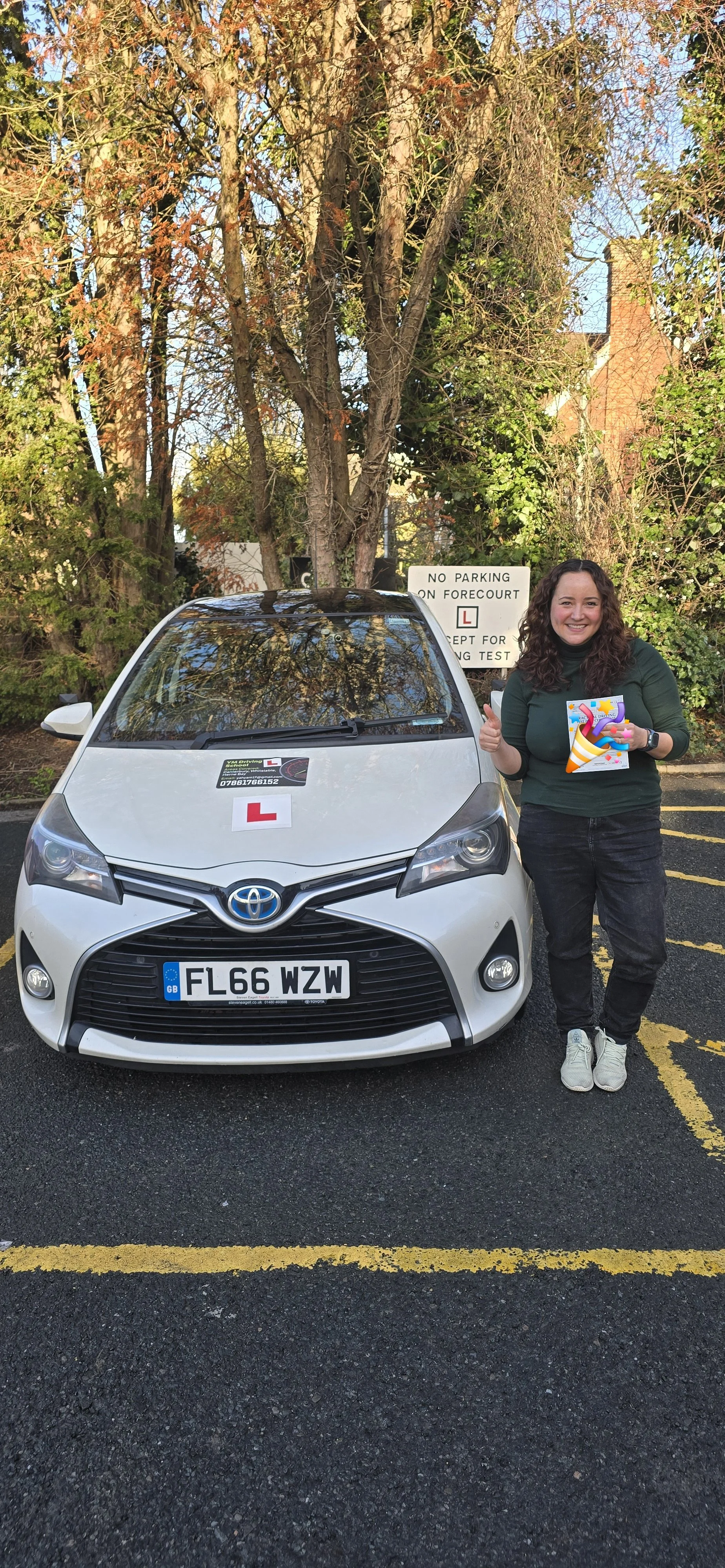 A young woman with curly hair holding a small colorful cake, giving a thumbs up next to a white driving school car with a learner's permit sign and a license plate reading FL66 WZW, parked in a lot with trees and a sign in the background.
