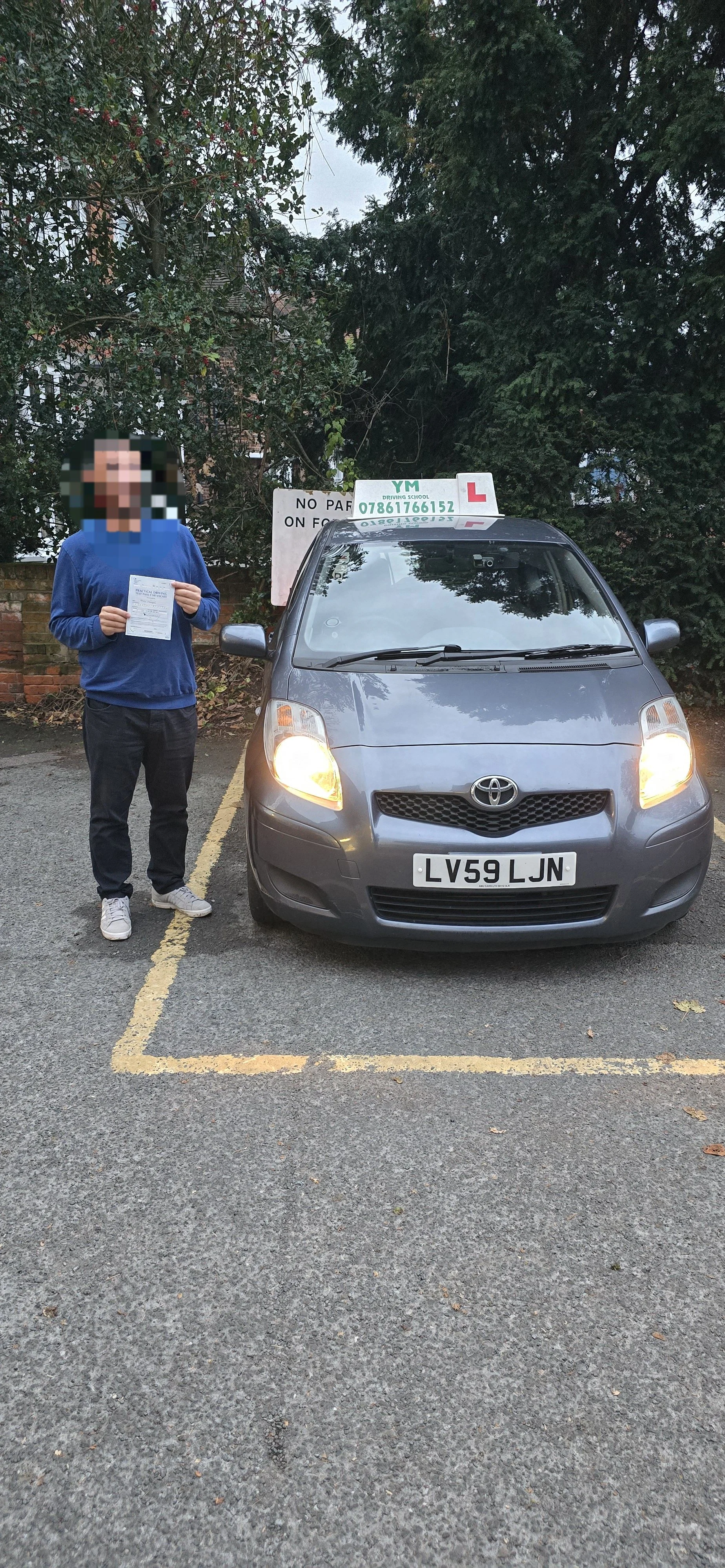 A person standing next to a gray Toyota car in a parking lot. The person is holding a paper, and the car has a driving school sign on top with a phone number and a learner's permit sign. The license plate reads LV59 LJN. There are trees and a 'No Par
