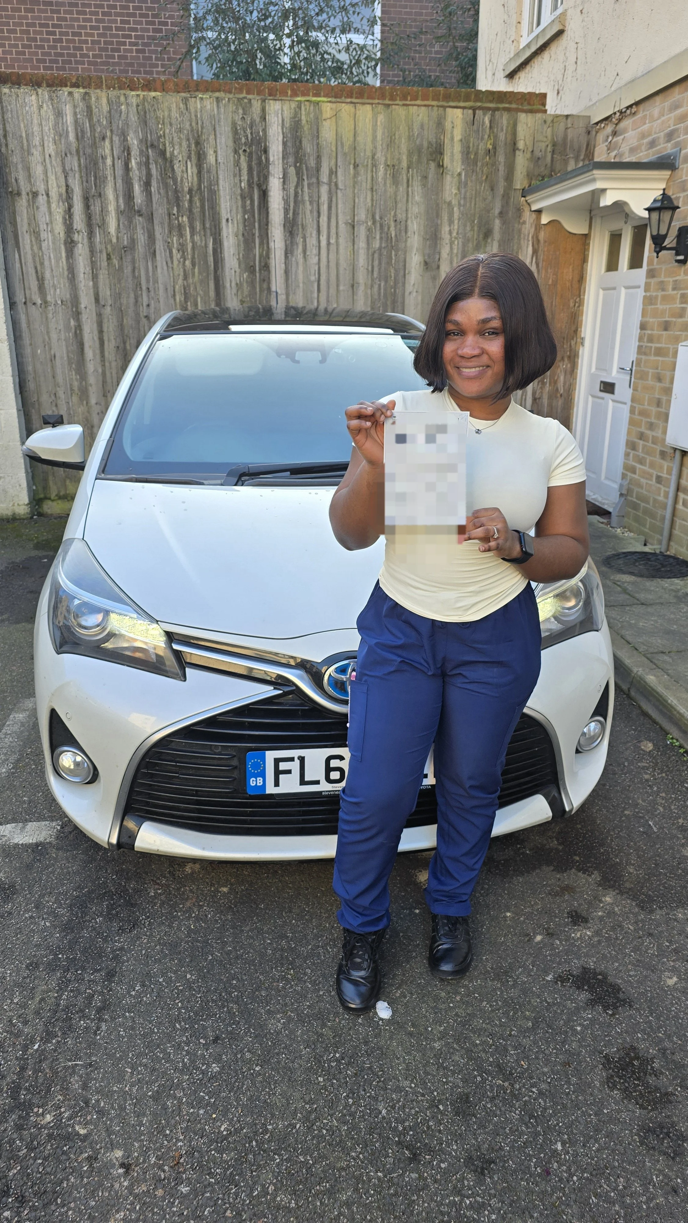A woman standing in front of a white Toyota car, holding a document, smiling, in a residential driveway.