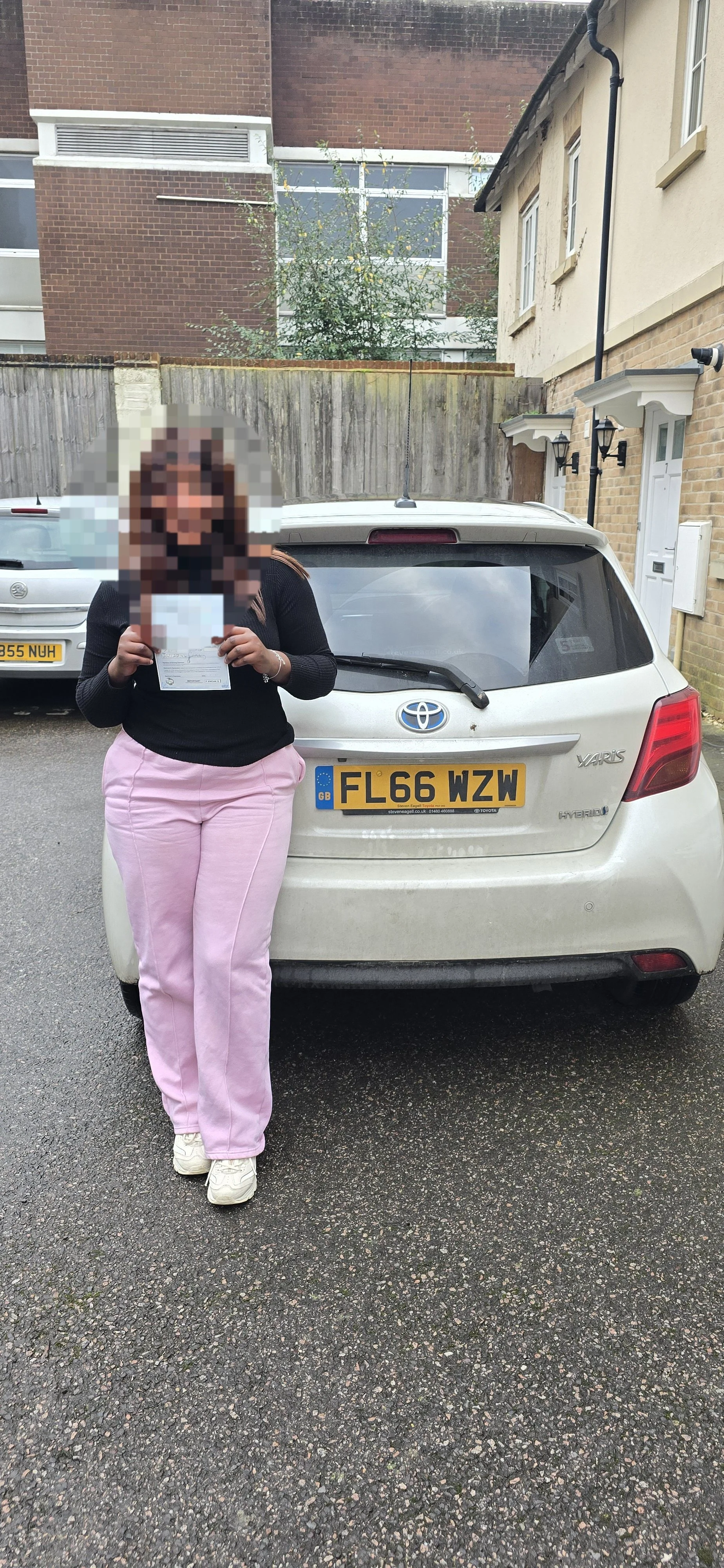 A woman with pixelated face holding a piece of paper, standing next to a white Toyota Yaris with a yellow UK license plate, in a parking lot with a brick and beige building in the background.