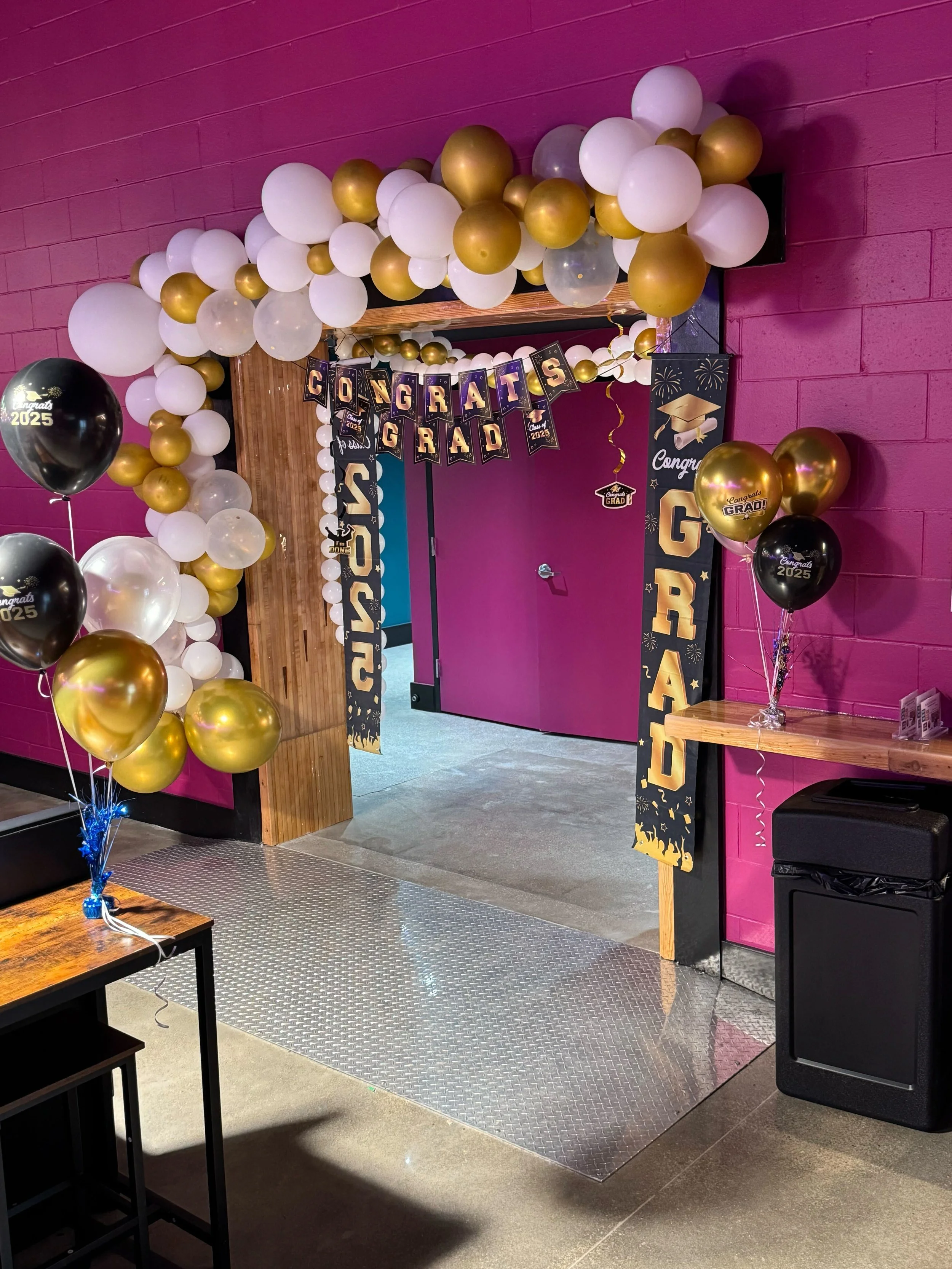 Decorated entrance with balloons and a banner celebrating graduation, featuring black, gold, and white balloons, and congratulatory signs.