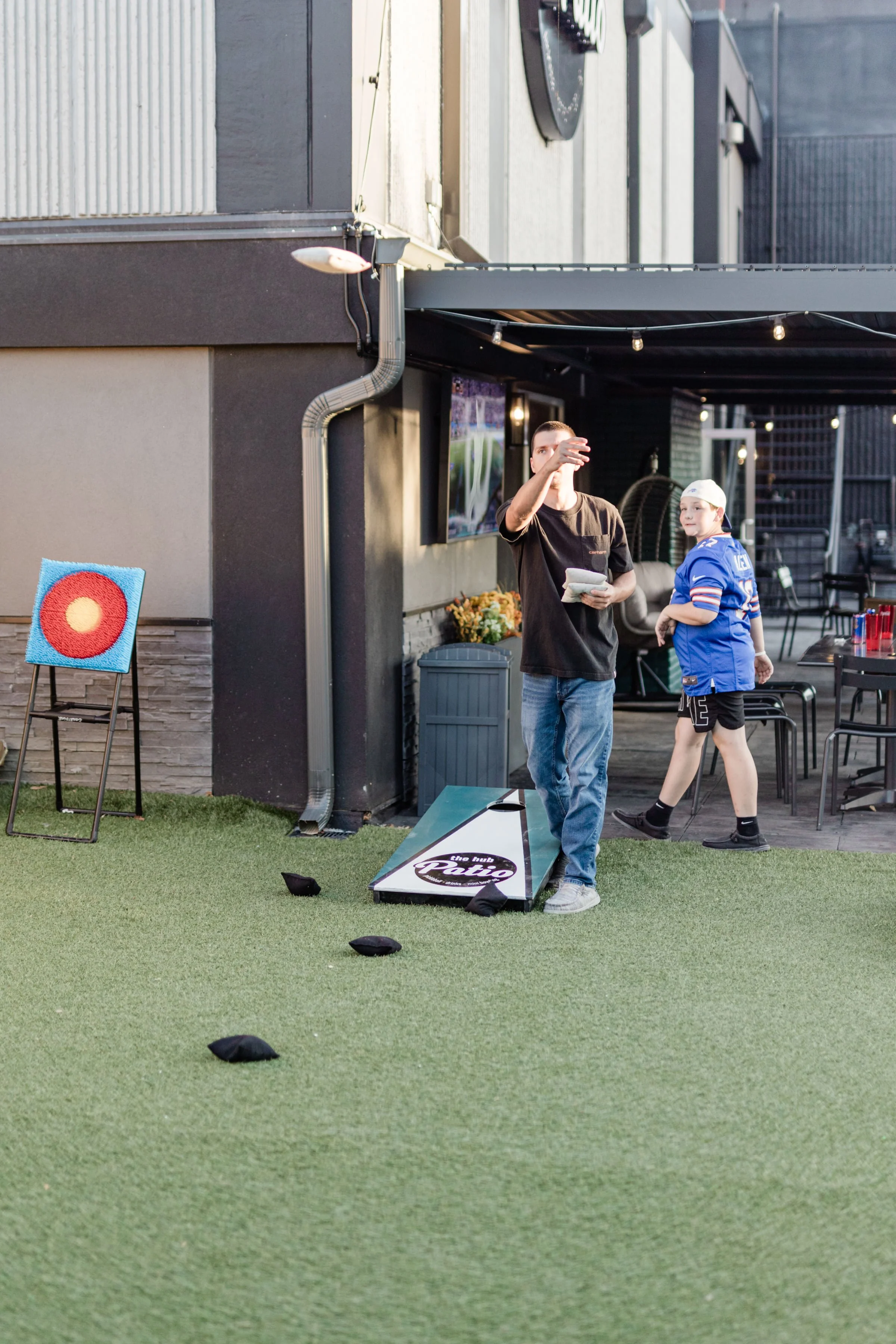 Two men playing outdoor cornhole near a bar or restaurant, one in a black t-shirt and jeans, the other in a blue sports jersey and shorts, with a small group of people visible in the background.