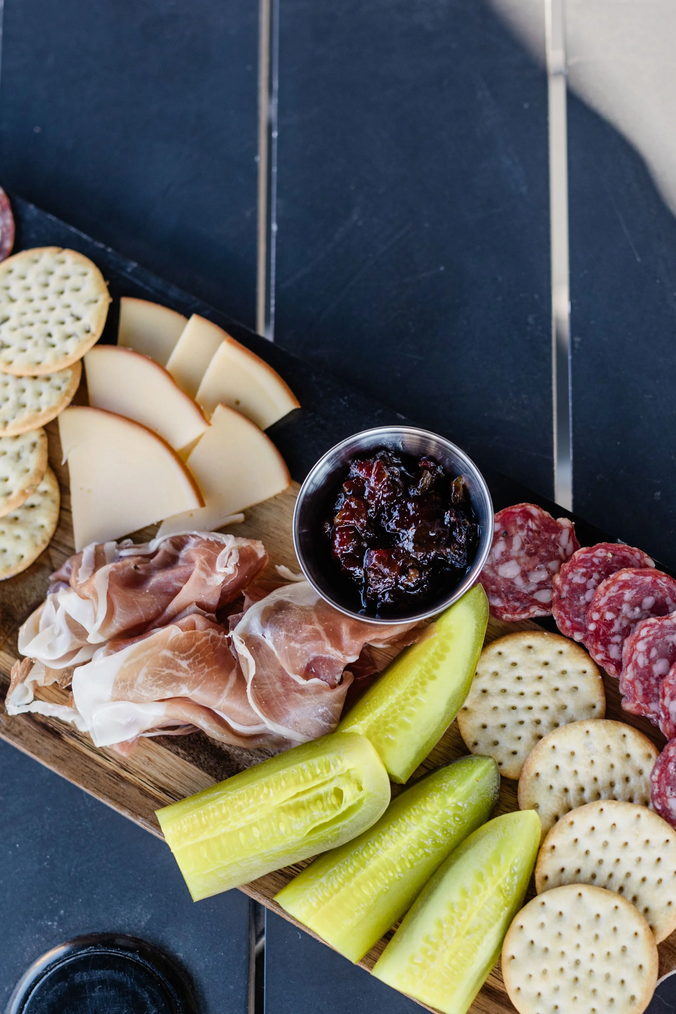 Charcuterie board with crackers, cheese, prosciutto, sliced pepperoni, sliced pickles, cranberry jam, and a glass of red wine.