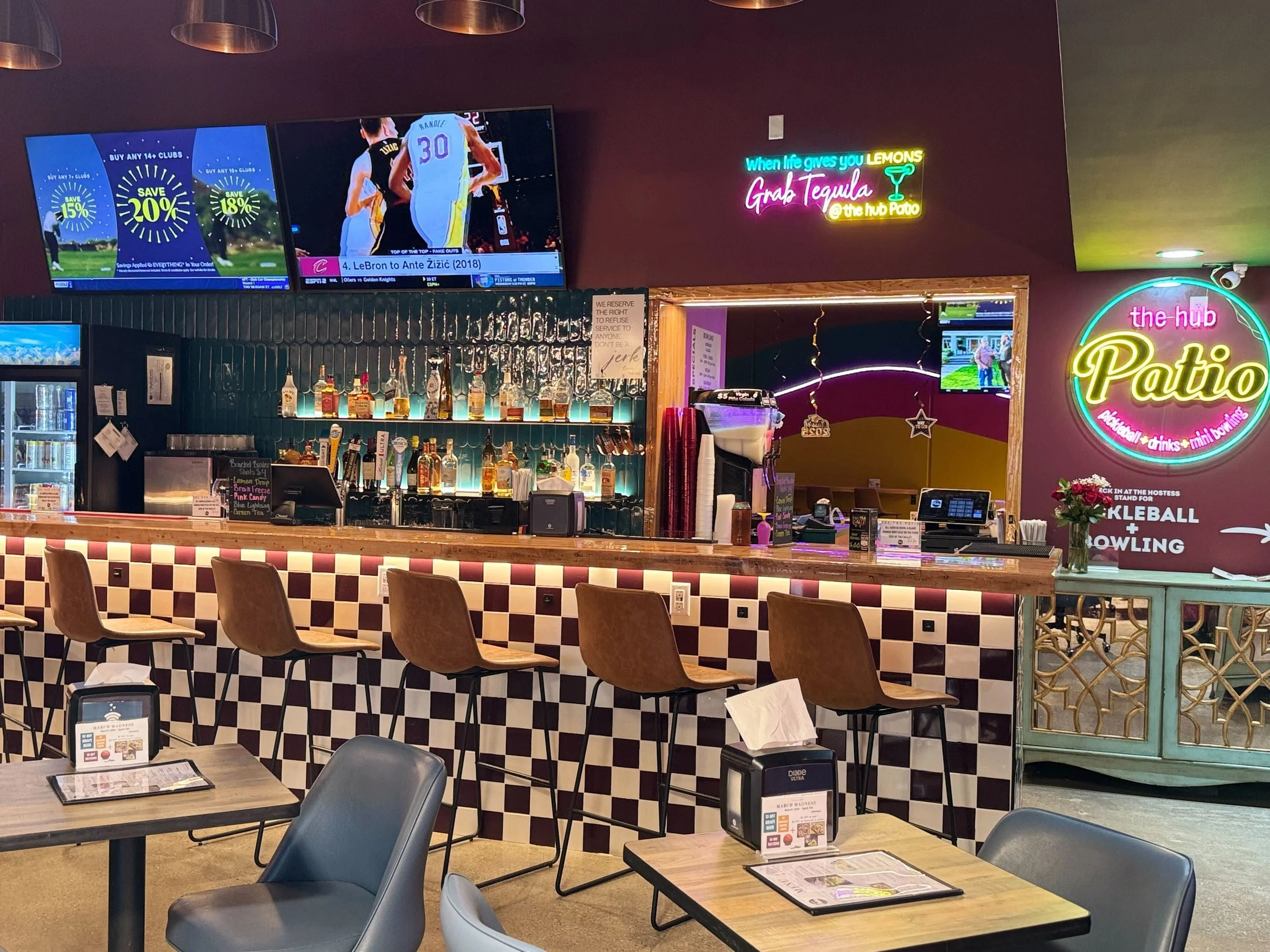 Interior of a bar with a checkered counter, bar stools, neon signs, a television showing a basketball game, and a seating area in the foreground.