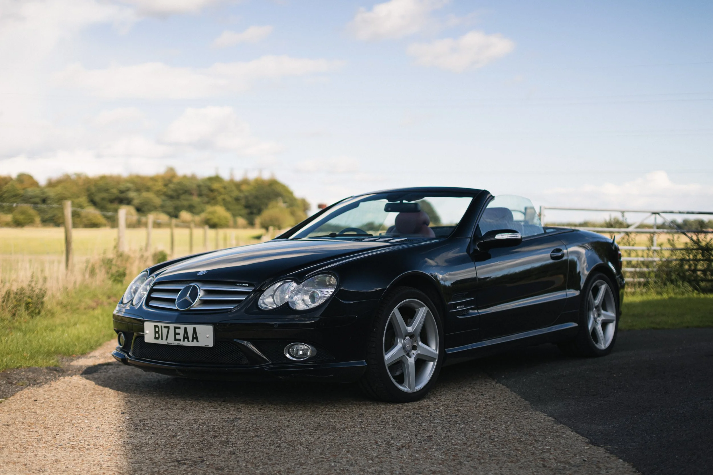 Black Mercedes-Benz convertible parked on the side of a rural road with green fields and trees in the background, under a partly cloudy sky.