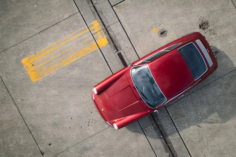 A top-down view of a red vintage Porsche parked on a concrete surface with yellow and black parking lot markings.