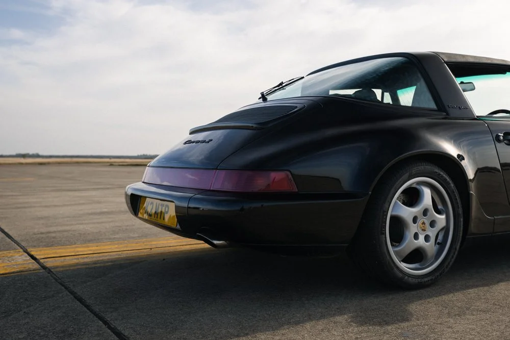 Black Porsche 911 convertible parked on an empty runway or tarmac with a cloudy sky in the background.