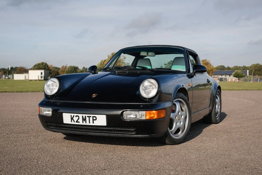 Black Porsche 911 sports car parked on an asphalt lot with grass and trees in the background.