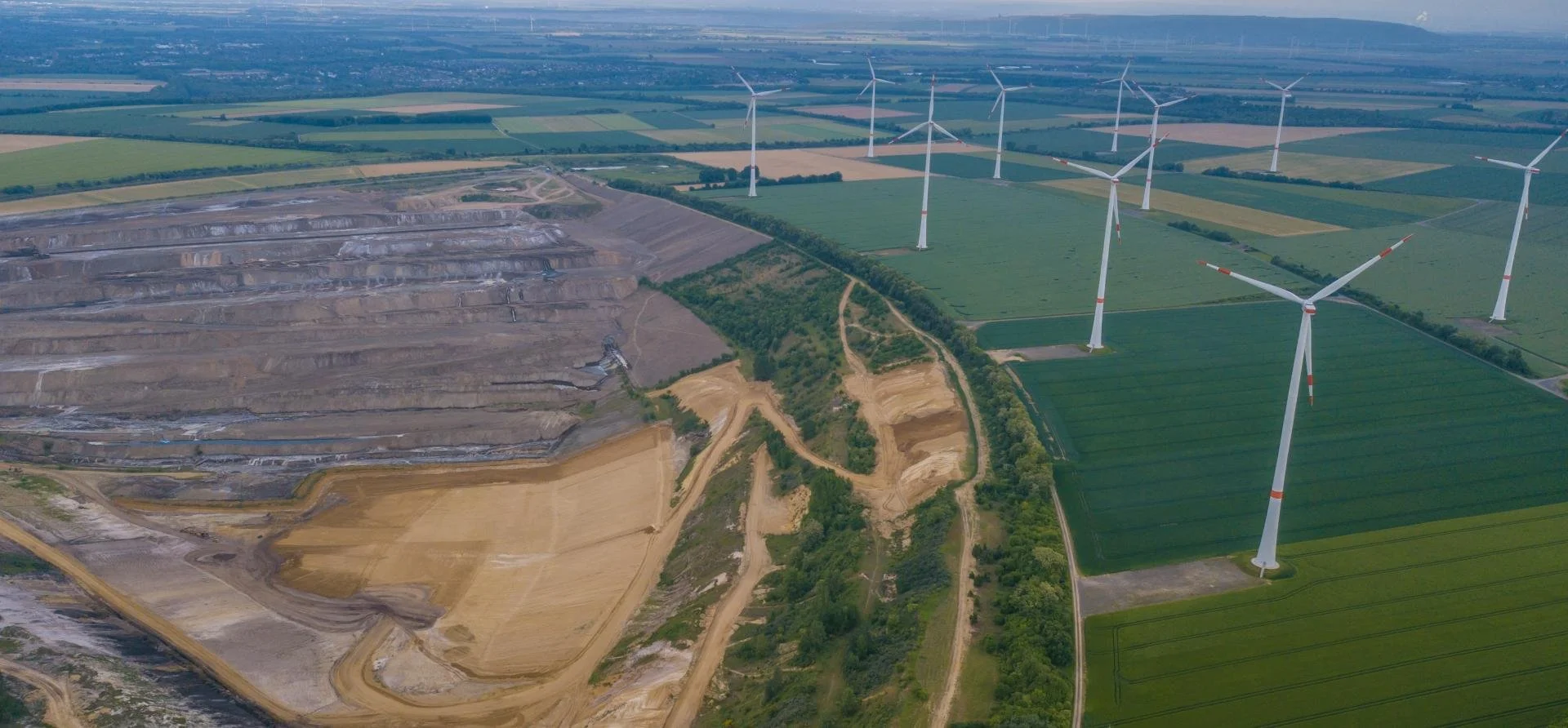 Aerial view of wind turbines in a green farmland area next to a brown construction or mining site with dirt roads and terraces.