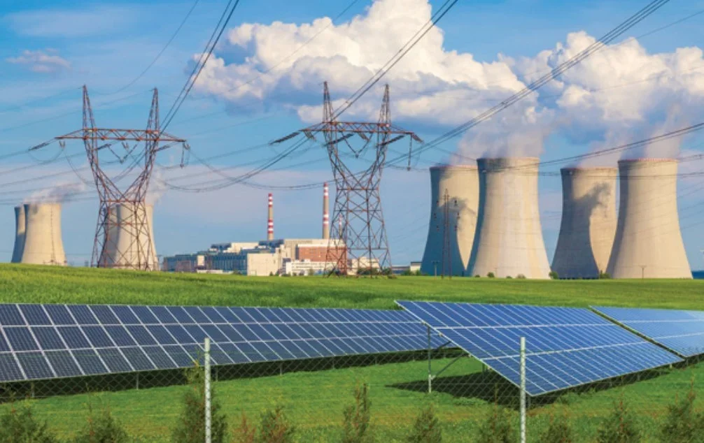 Solar panels in the foreground with a nuclear power plant and cooling towers in the background, under a partly cloudy sky.
