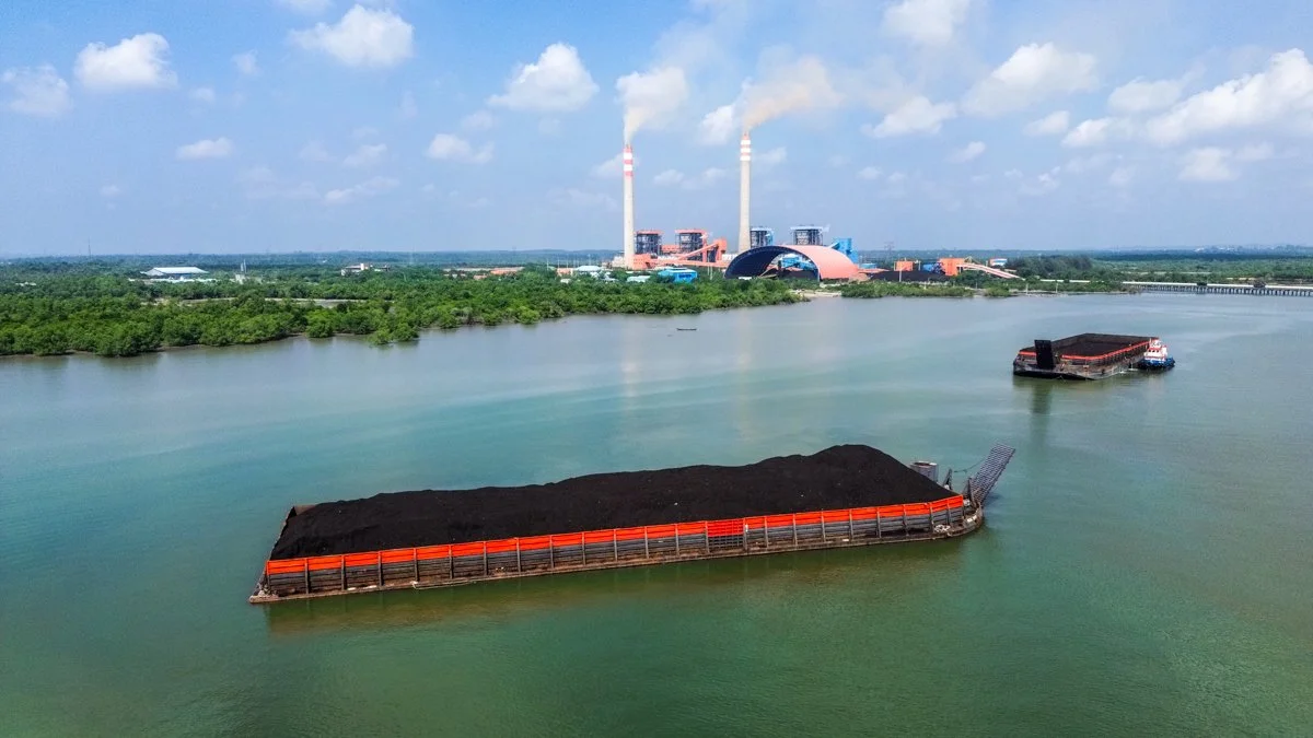 Two barges filled with coal floating on a body of water with an industrial power plant in the background.