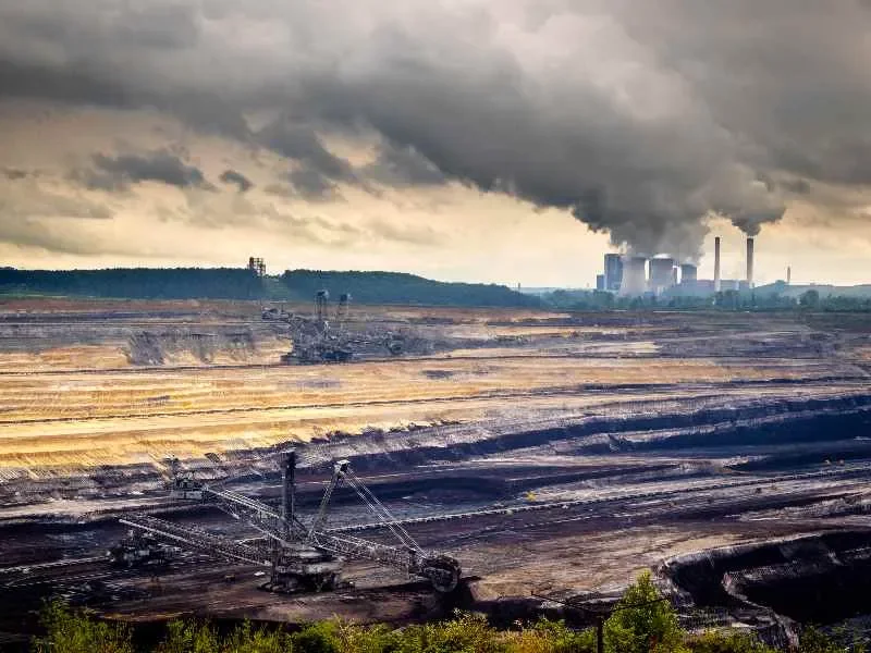 An open-pit coal mine with large excavators working and smoke billowing from distant power plant smokestacks under a cloudy sky.