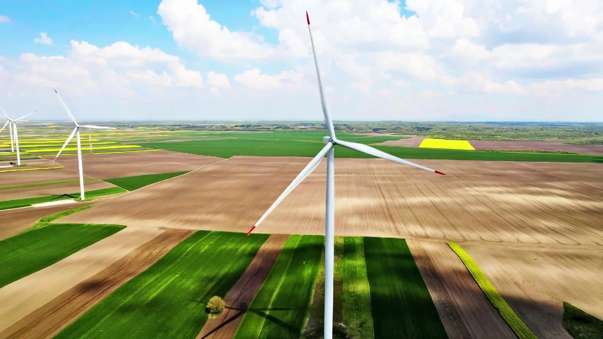 A large wind turbine stands in a green and brown patchwork field under a partly cloudy sky.