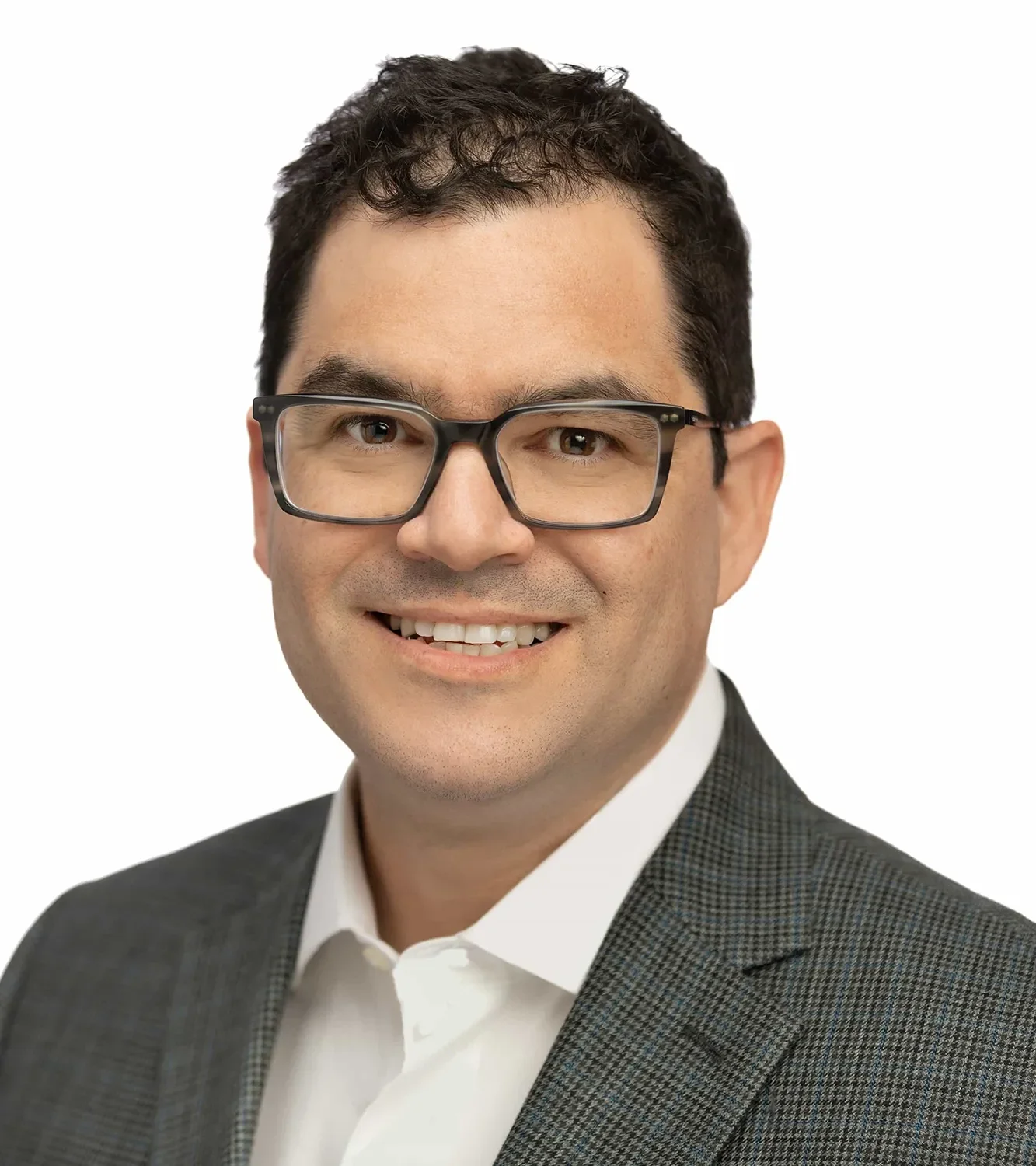 Close-up of a man with dark hair, wearing glasses, smiling, dressed in a dark checked suit and white shirt, against a white background.