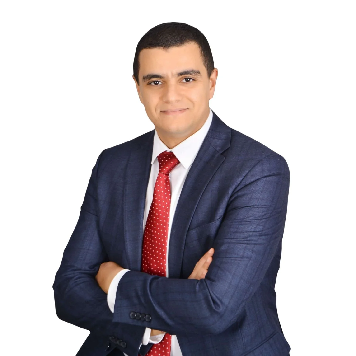 A young man in a dark blue suit with a white shirt and a red polka dot tie, standing with arms crossed against a plain white background.