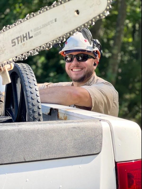 Owner & Operator, Duncan Gilbert, wearing a protective helmet and sunglasses smiling while working with a chainsaw aside of his company truck. Red Line Tree Care.
