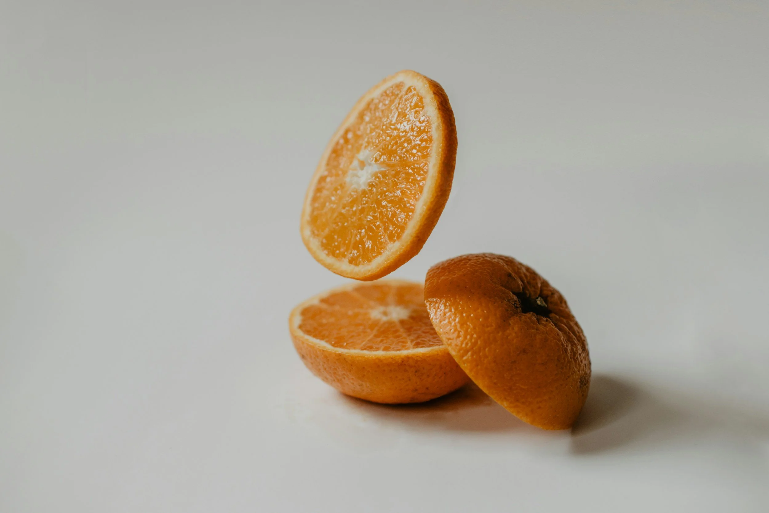 A peeled orange with a slice suspended mid-air above a halved orange on a plain white background.