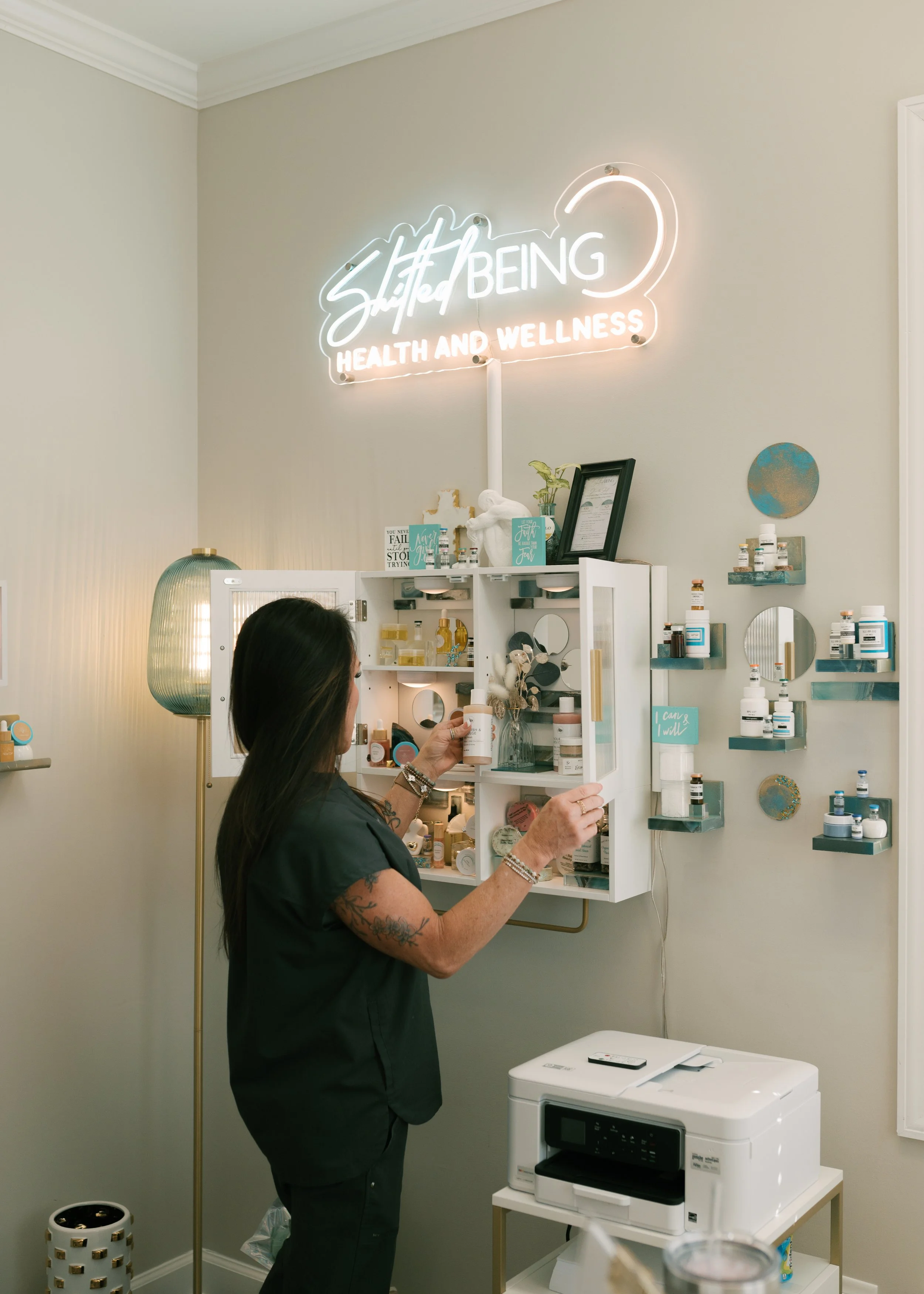 A woman with tattoos shopping for health and wellness products in a modern, well-lit retail space with a neon sign above that reads 'Still and Being Health and Wellness'. She is holding a bottle and standing in front of a white wall-mounted display cabinet filled with various bottles and containers.