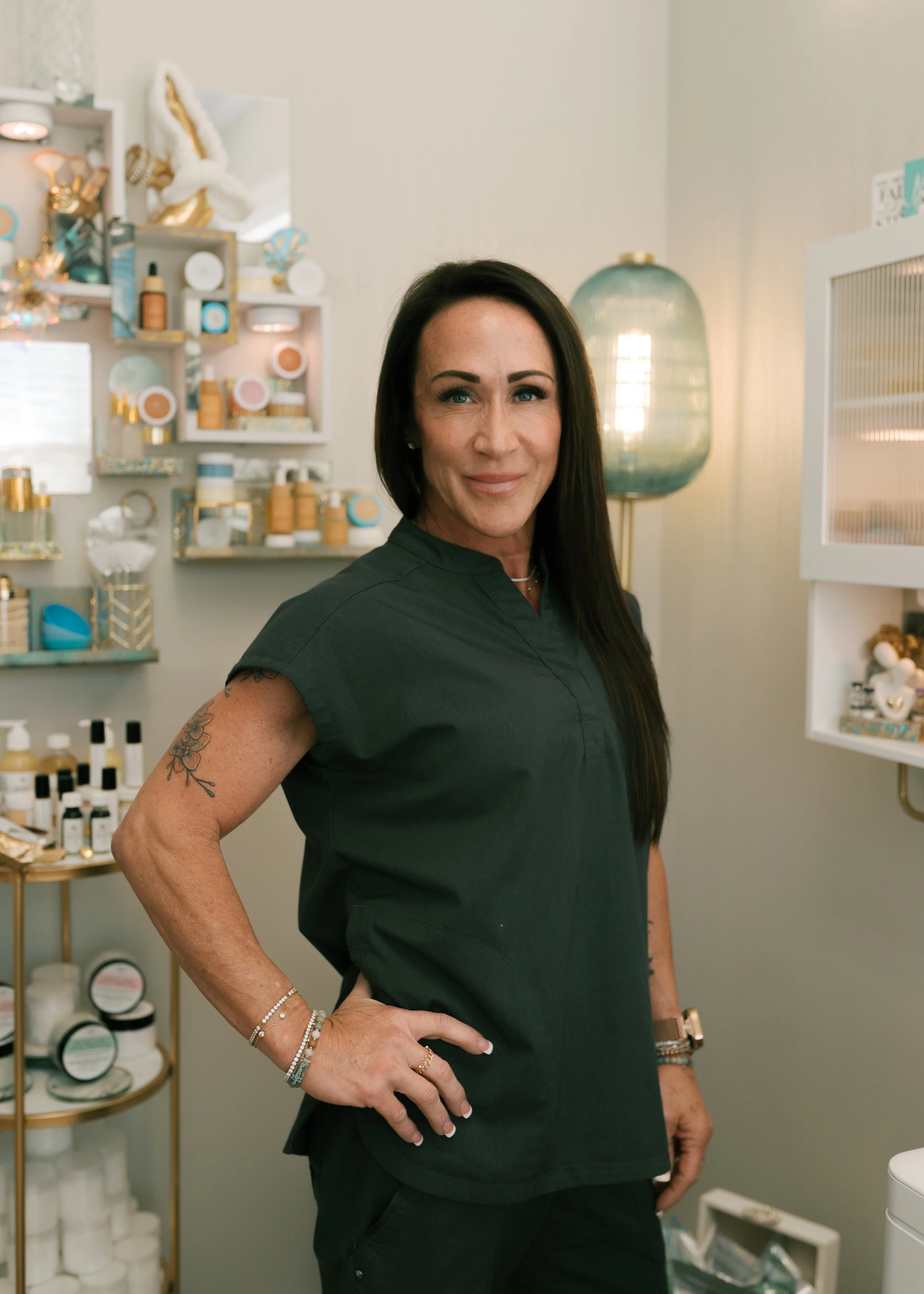 A woman with long dark hair in a dark scrub top standing in a boutique or spa with skincare products on shelves behind her.