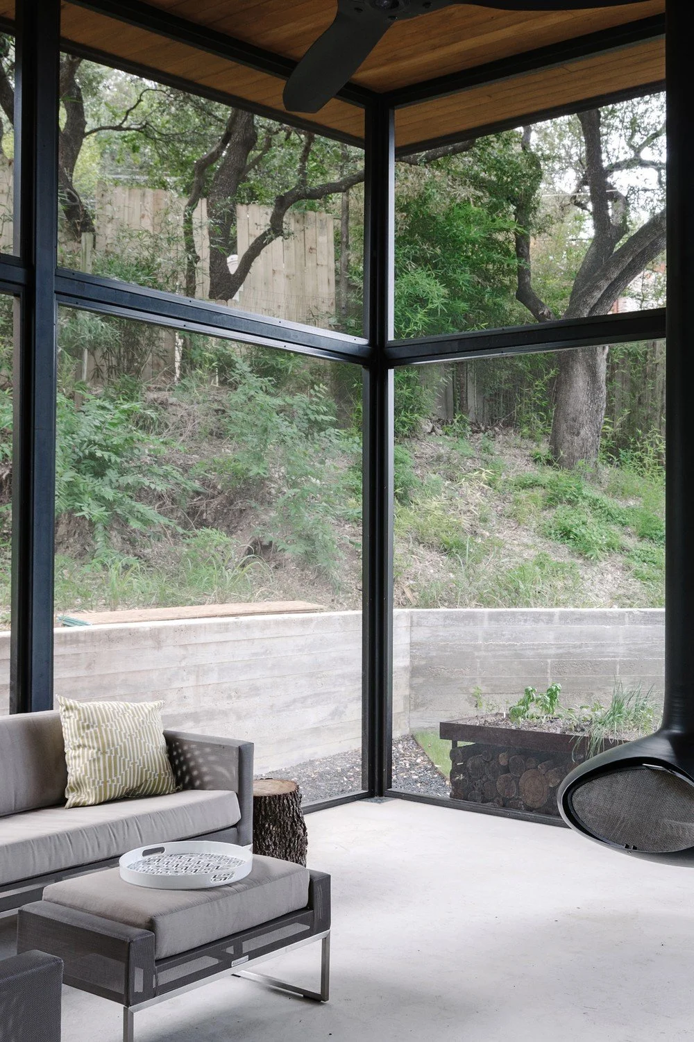 Rockingham Residence by Forsite Studio — screened porch with black metal framing, wood ceiling, gray sofa and a suspended fireplace, open to oak woodland — Austin, TX