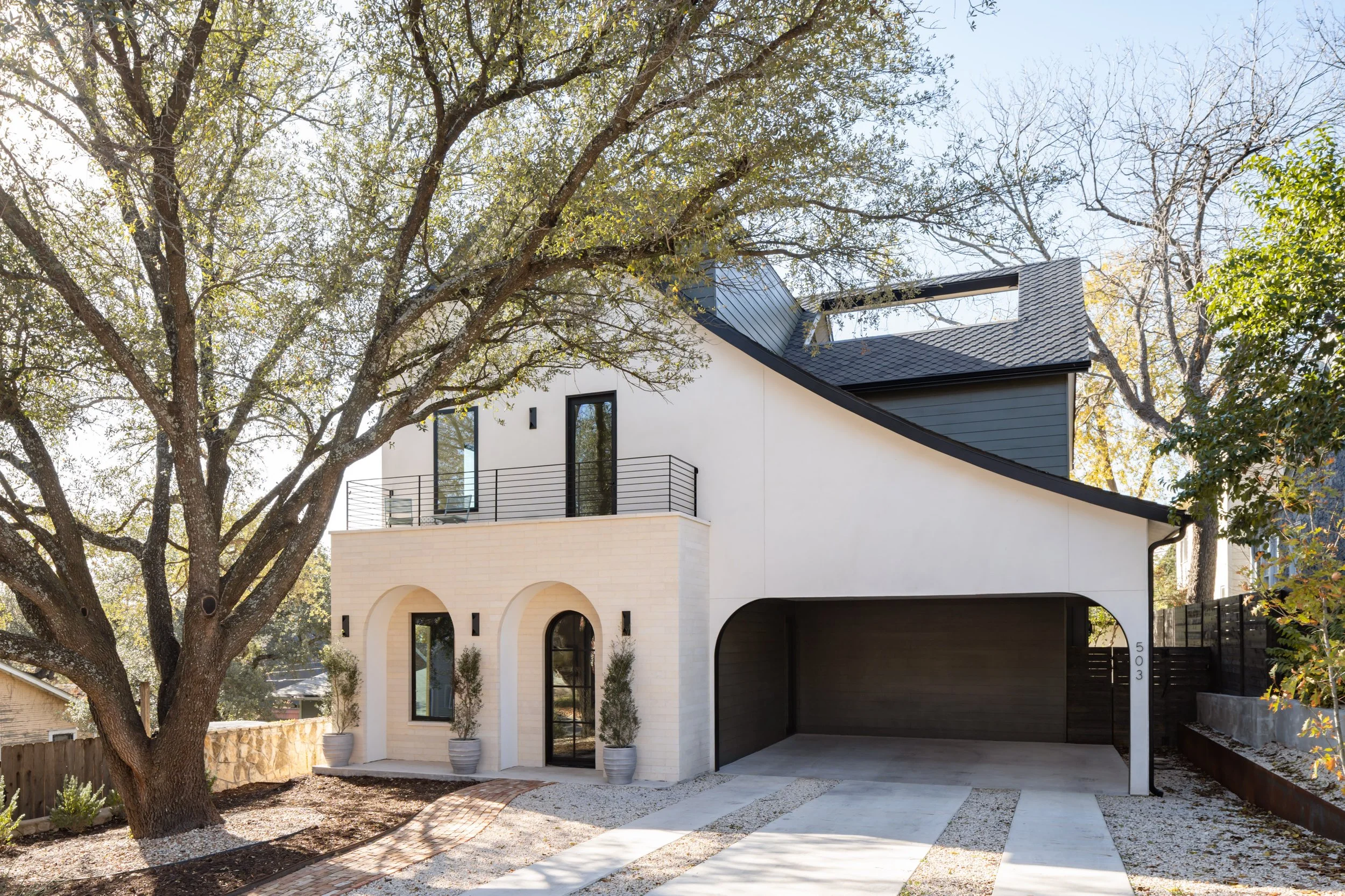 Modern two-story house with white and dark gray exterior walls, arched windows, a small balcony, and a garage. Large tree in front with potted plants near the entrance.