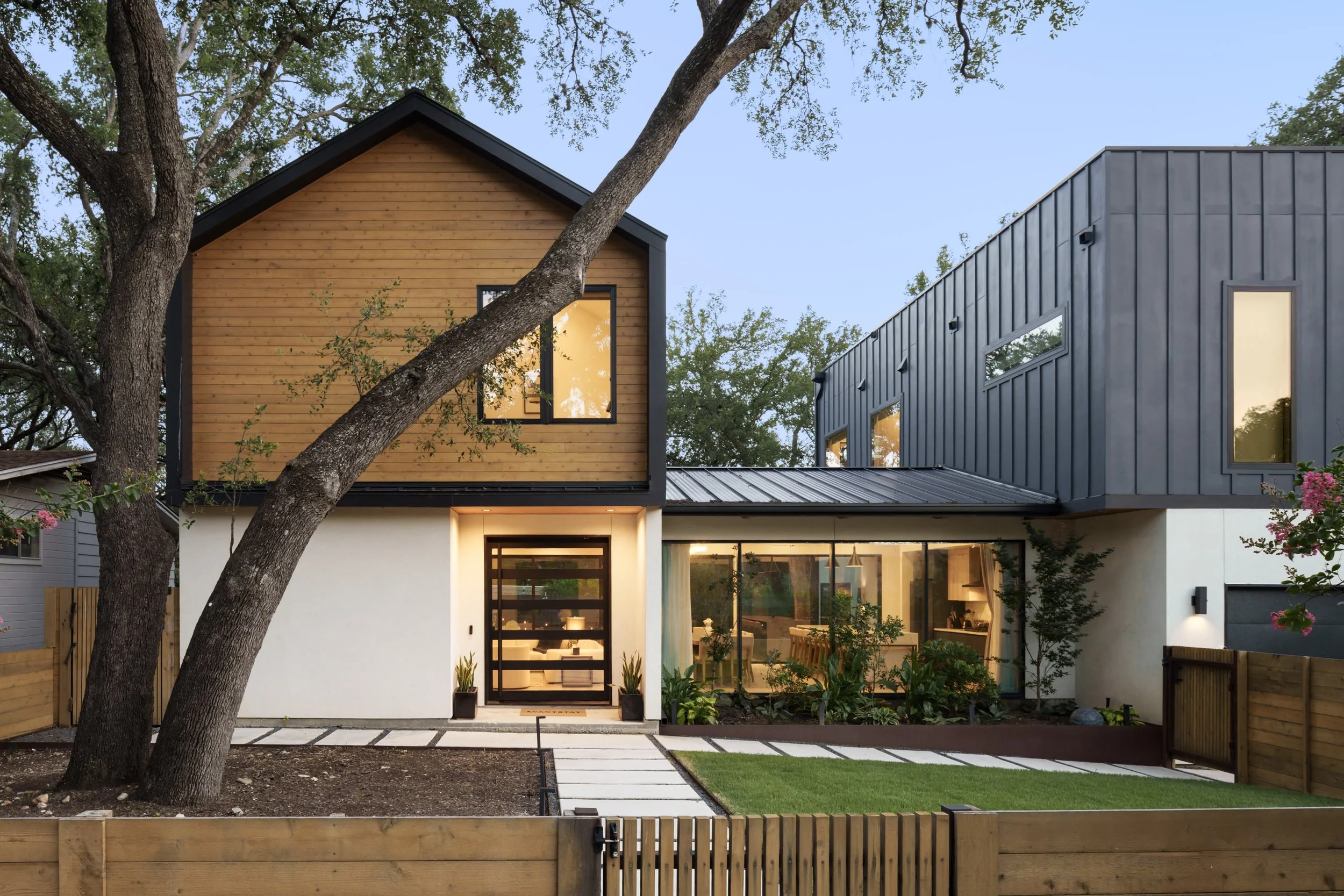 Mariposa Residence by Forsite Studio — glowing entry at
dusk beneath a cedar gable, with a live oak leaning across the front of
 the house — Austin, TX