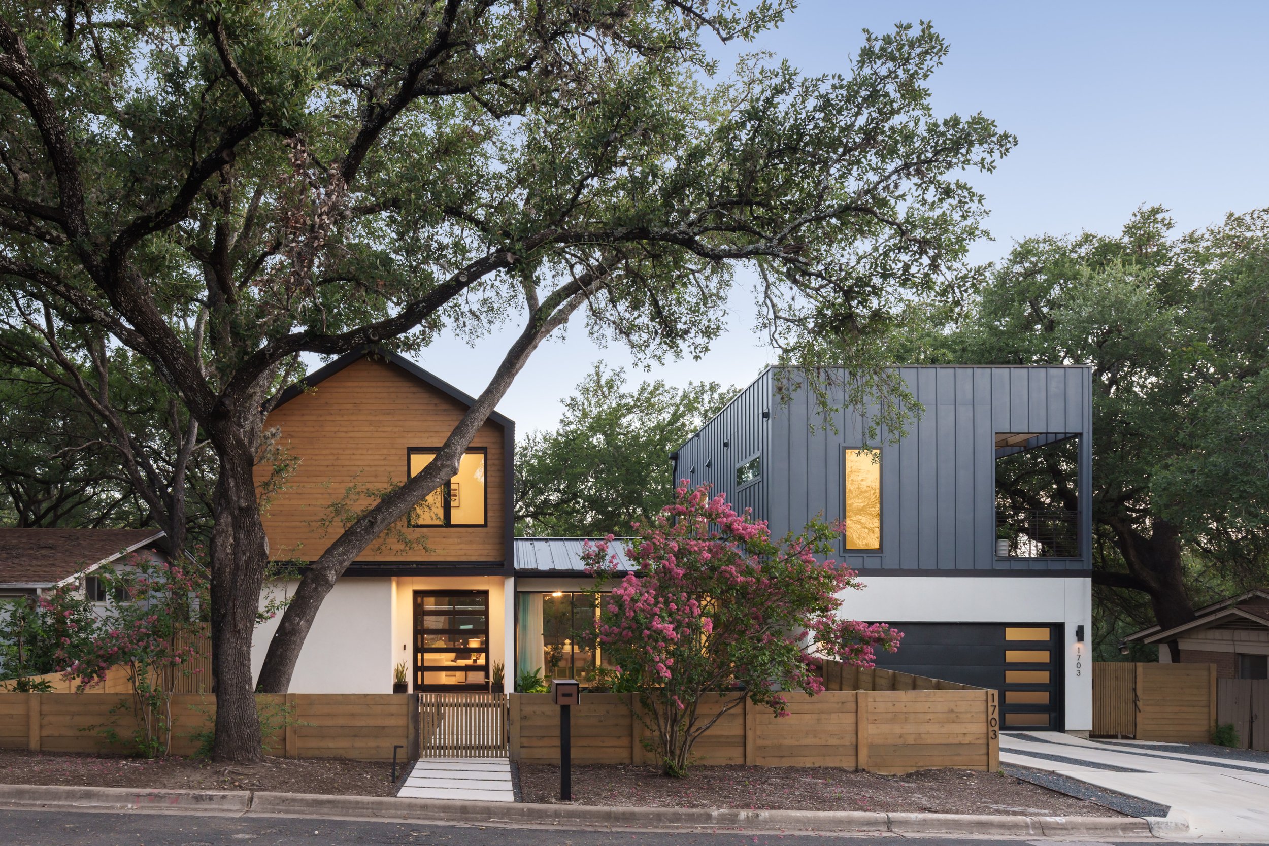 Mariposa Residence by Forsite Studio — glowing entry at
dusk beneath a cedar gable, with a live oak leaning across the front of
 the house — Austin, TX