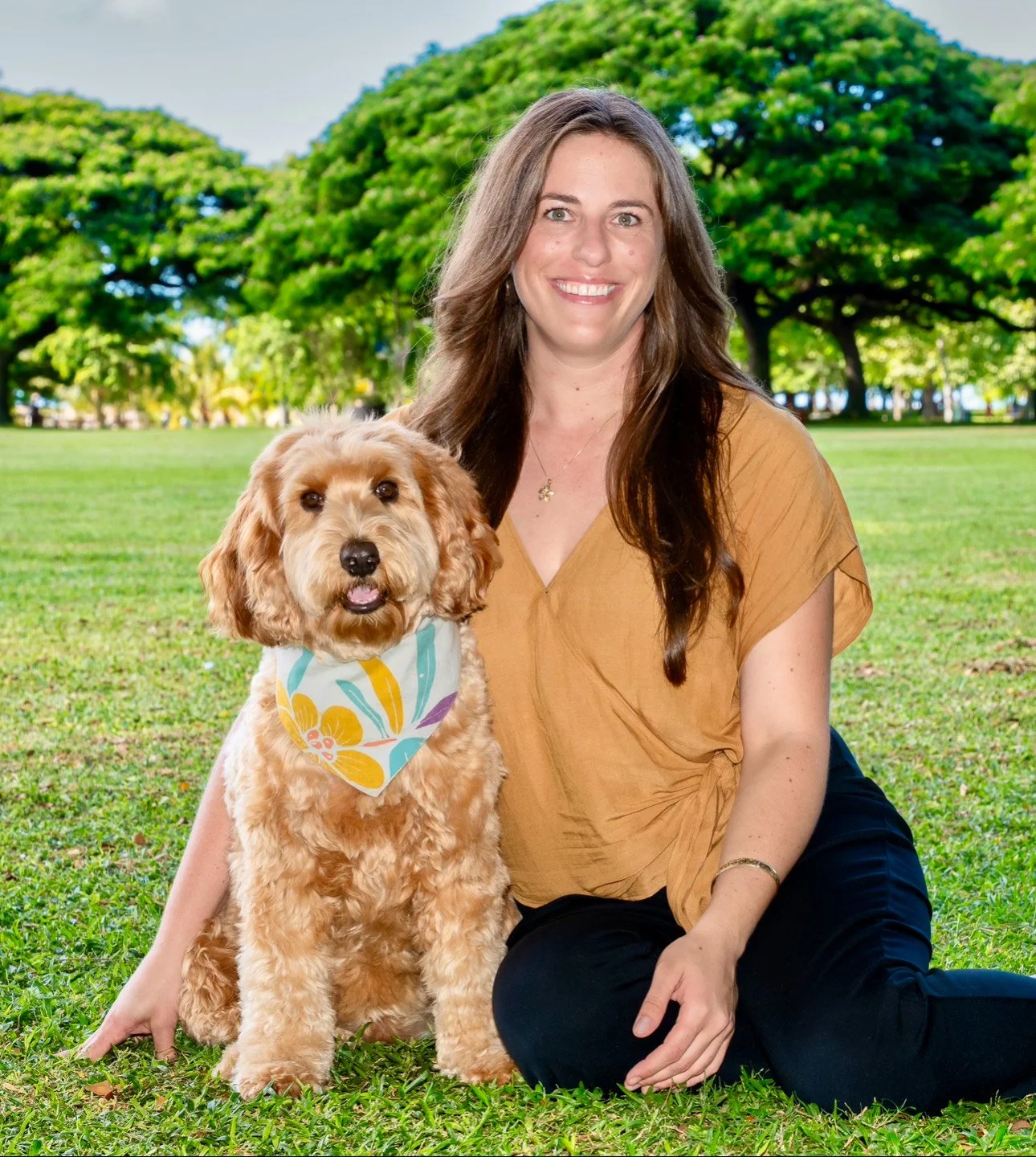 A woman with long brown hair, wearing a tan top and black pants, sits on grass in a park next to a golden retriever wearing a colorful bandana. They are smiling and surrounded by green trees.