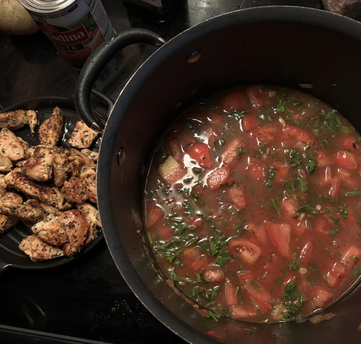A pot of homemade tomato and vegetable soup next to a plate of cooked, seasoned chicken pieces.