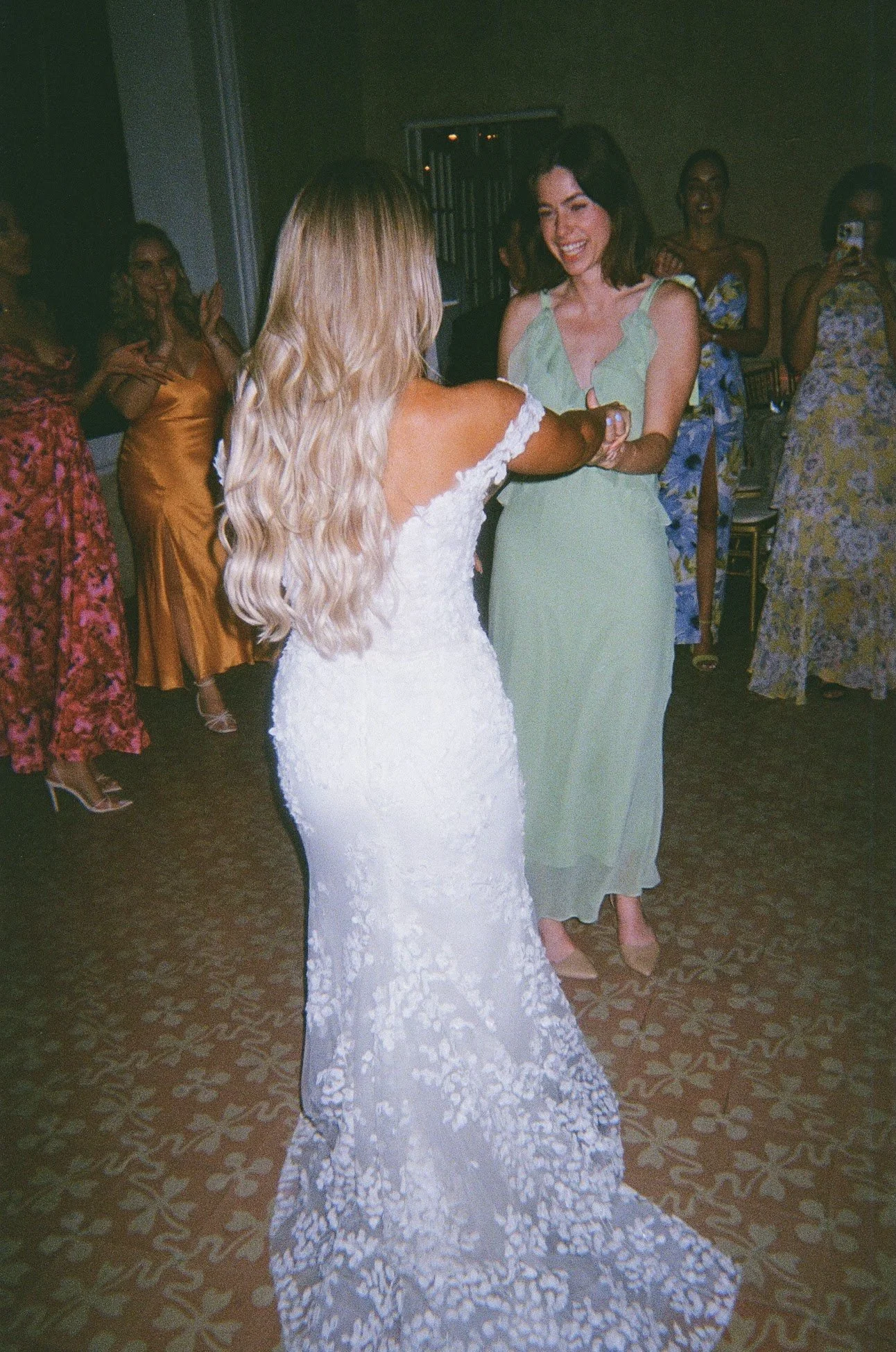 Two women dancing at a celebration, with the woman in a white lace gown and the other in a light green dress, surrounded by guests in colorful dresses.
