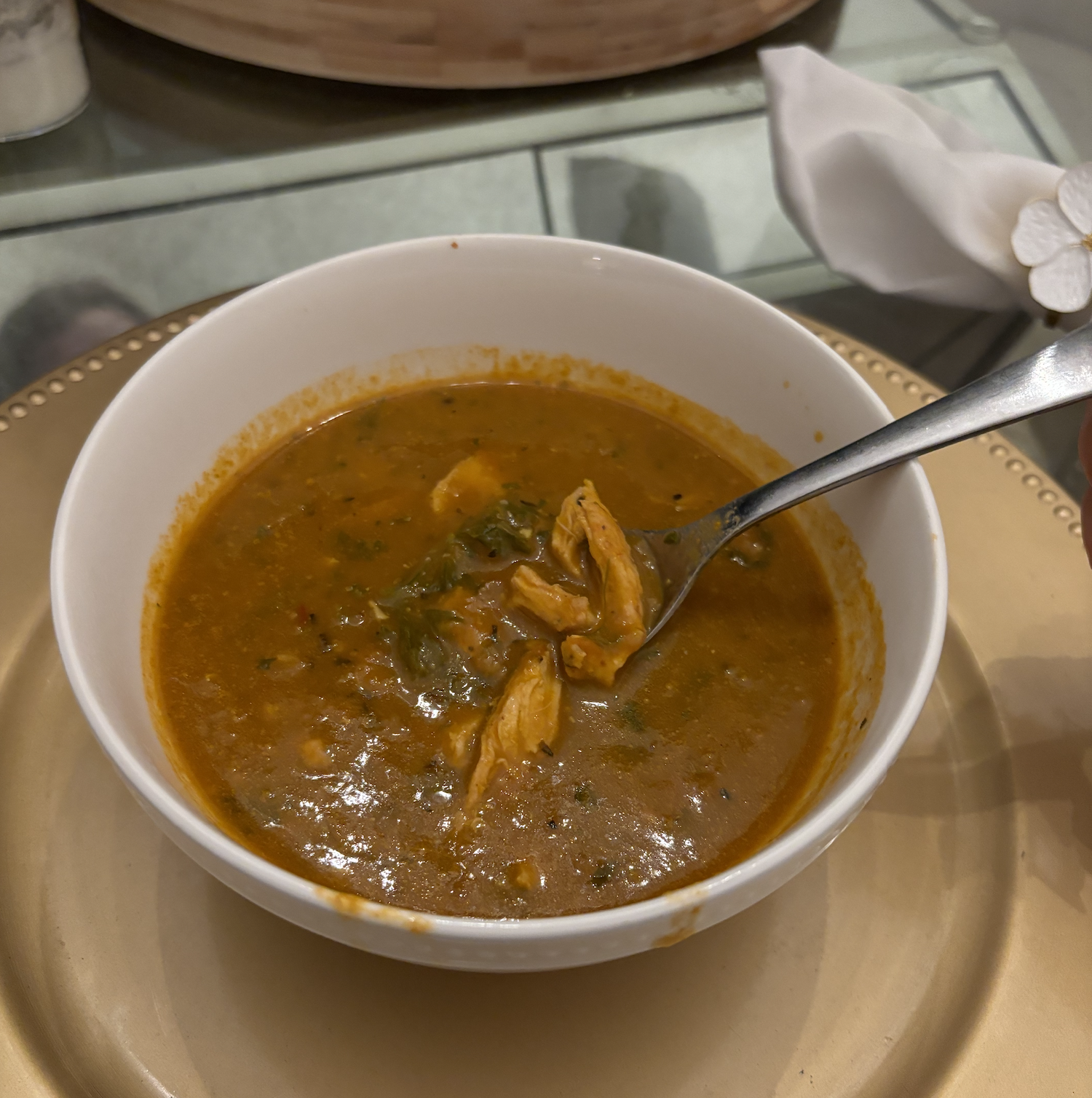 A white bowl filled with a thick, brownish-orange soup containing shredded chicken and greens, placed on a beige tray on a glass table.