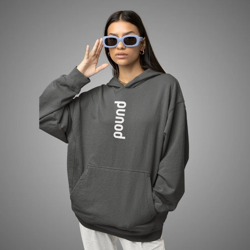 Woman with brown hair in a ponytail, wearing Space Gray POUND Hoodie with blue goth style text and a damnum button jean, standing against a plain gray wall.