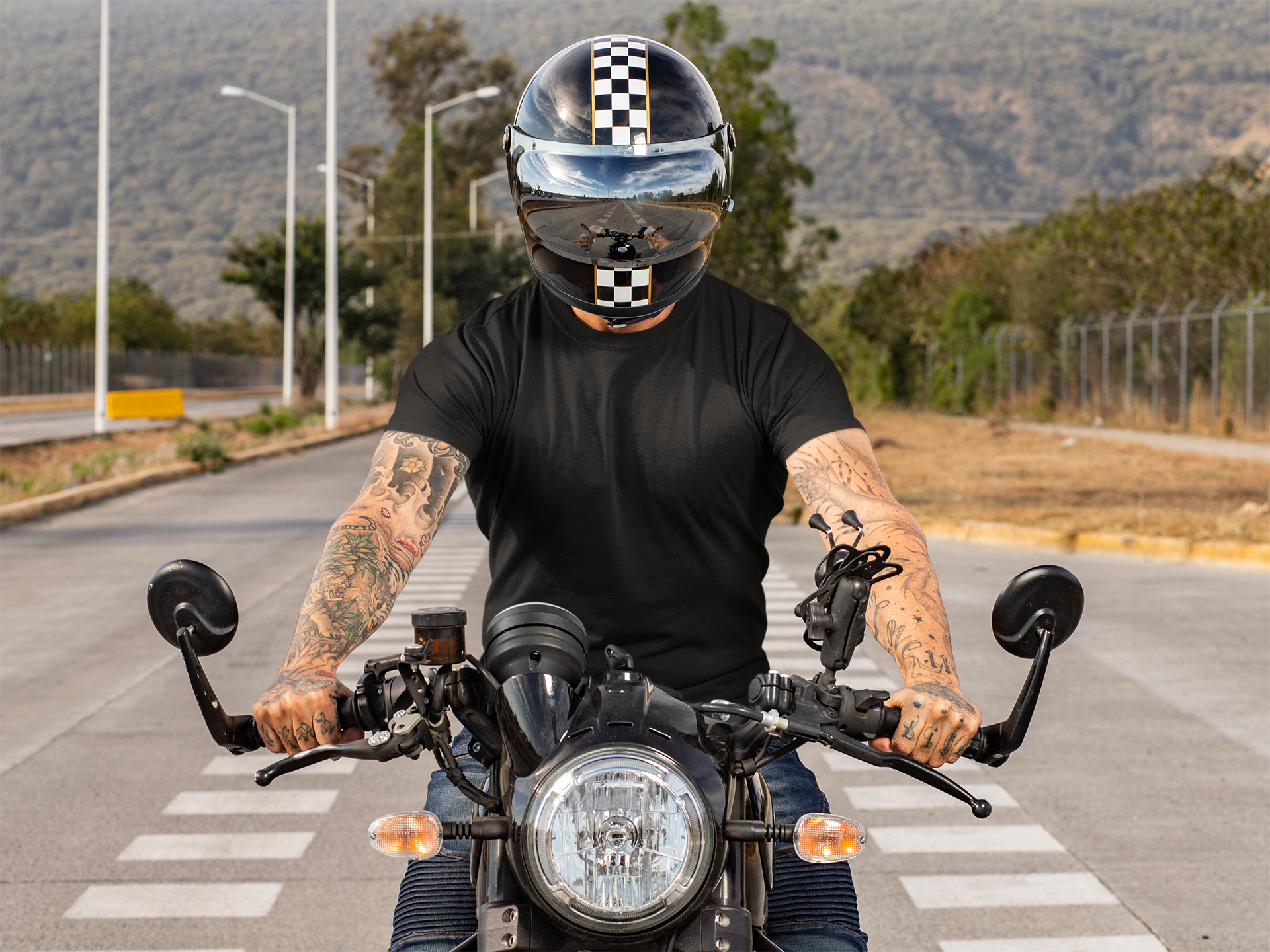 Man wearing a black shirt and a helmet with a checkered stripe, riding a motorcycle on a crosswalk on a street with trees and hills in the background.