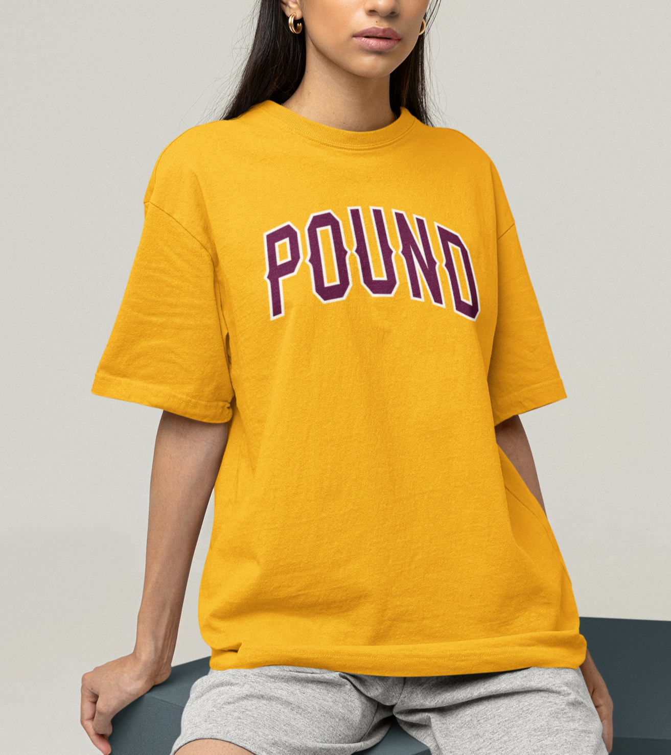 Woman wearing a t-shirt with POUND  across the center, a garment-washed heavyweight cotton POUND, bold lettering in burgundy with light gray accent trim. 6.0oz fabric 100% Cotton