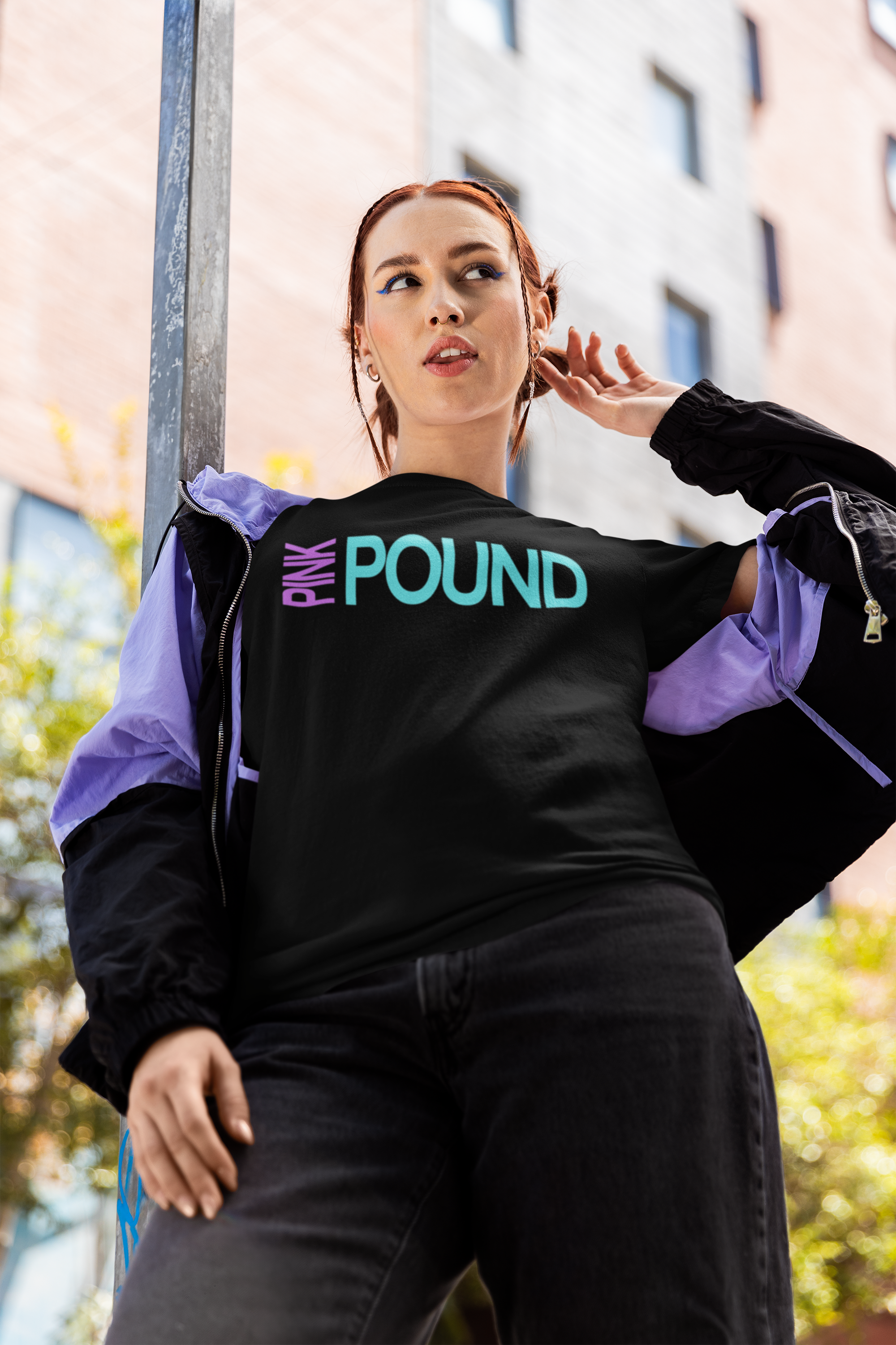 Young woman with red hair wearing a black t-shirt with colorful text, purple and black windbreaker, standing outdoors near a building and a pole, looking to the side.
