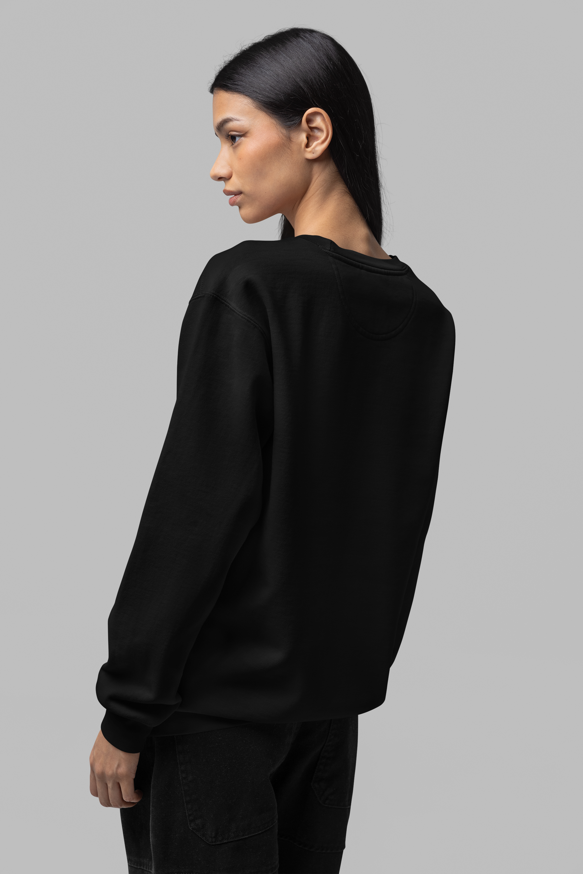 A woman with long black hair wearing a black sweatshirt and black jeans, standing against a plain gray background. She is shown in profile, with her face turned to the left and her left arm hanging down.