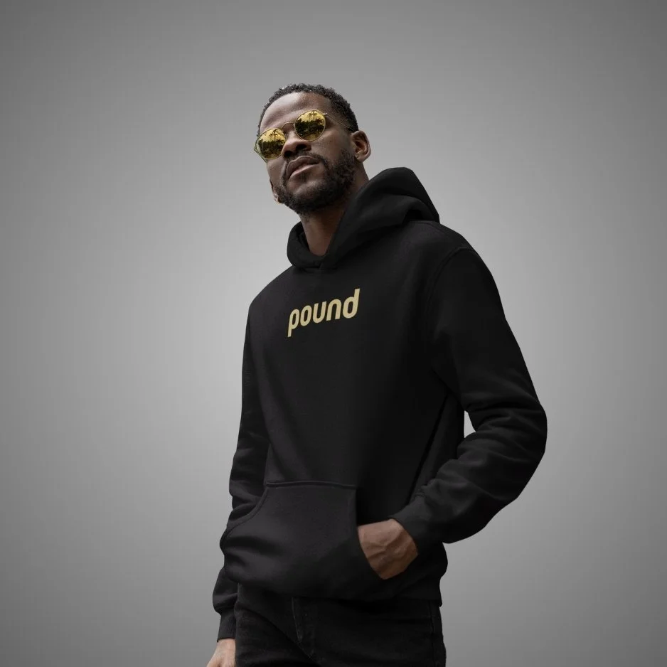 A man wearing reflective sunglasses and a black hoodie with the word 'pound' printed on it, standing against a plain gray background with hands in pockets.