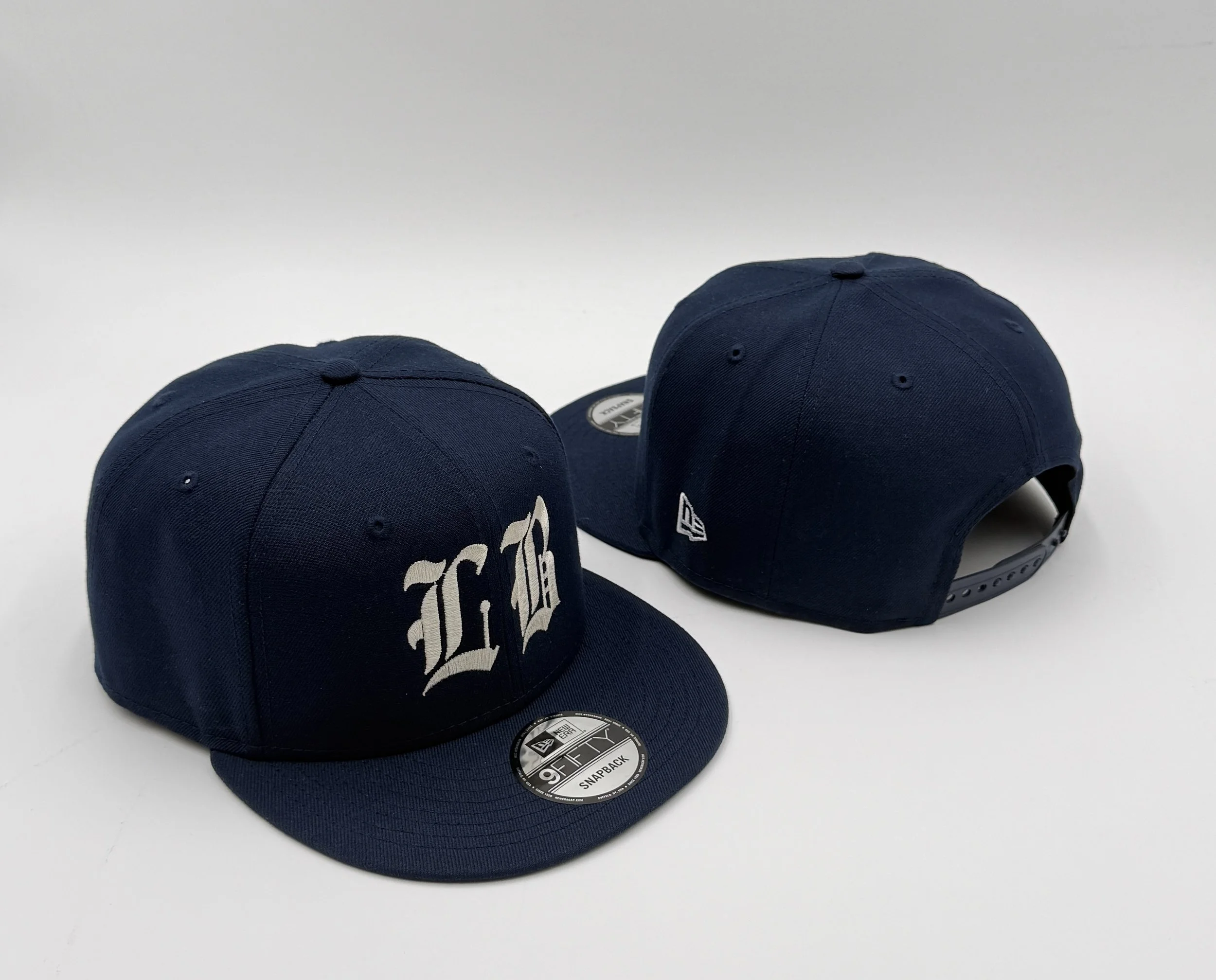 Three navy blue baseball caps on a plain white surface, with one cap showing the logo and two caps with visible stickers on the brim.