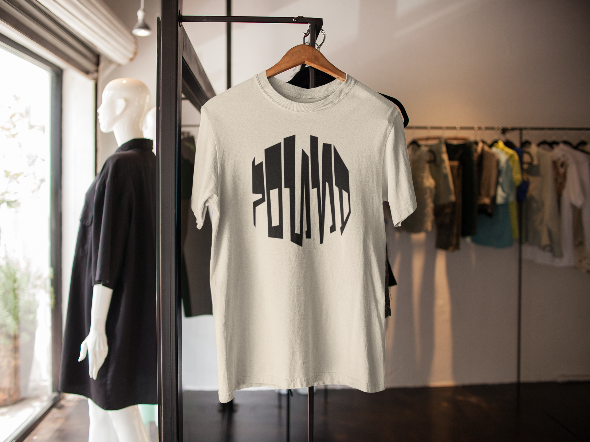 A white T-shirt hanging on a wooden hanger displays the word "DEAD END" in black, stylized, vertical text. The T-shirt is in a clothing store, with a mannequin dressed in black clothing nearby and various other garments hanging on racks in the background.