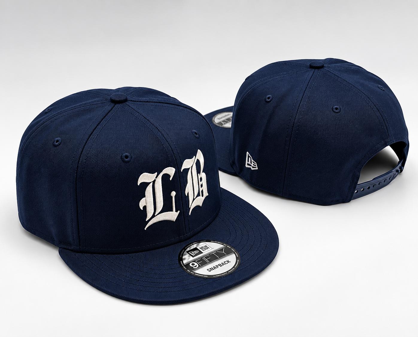 Two navy blue baseball caps, one with a white embroidered logo and the other with a small white logo, placed on a white surface. The cap in the foreground has a sticker on the brim indicating it is a New Era 9FIFTY snapback.