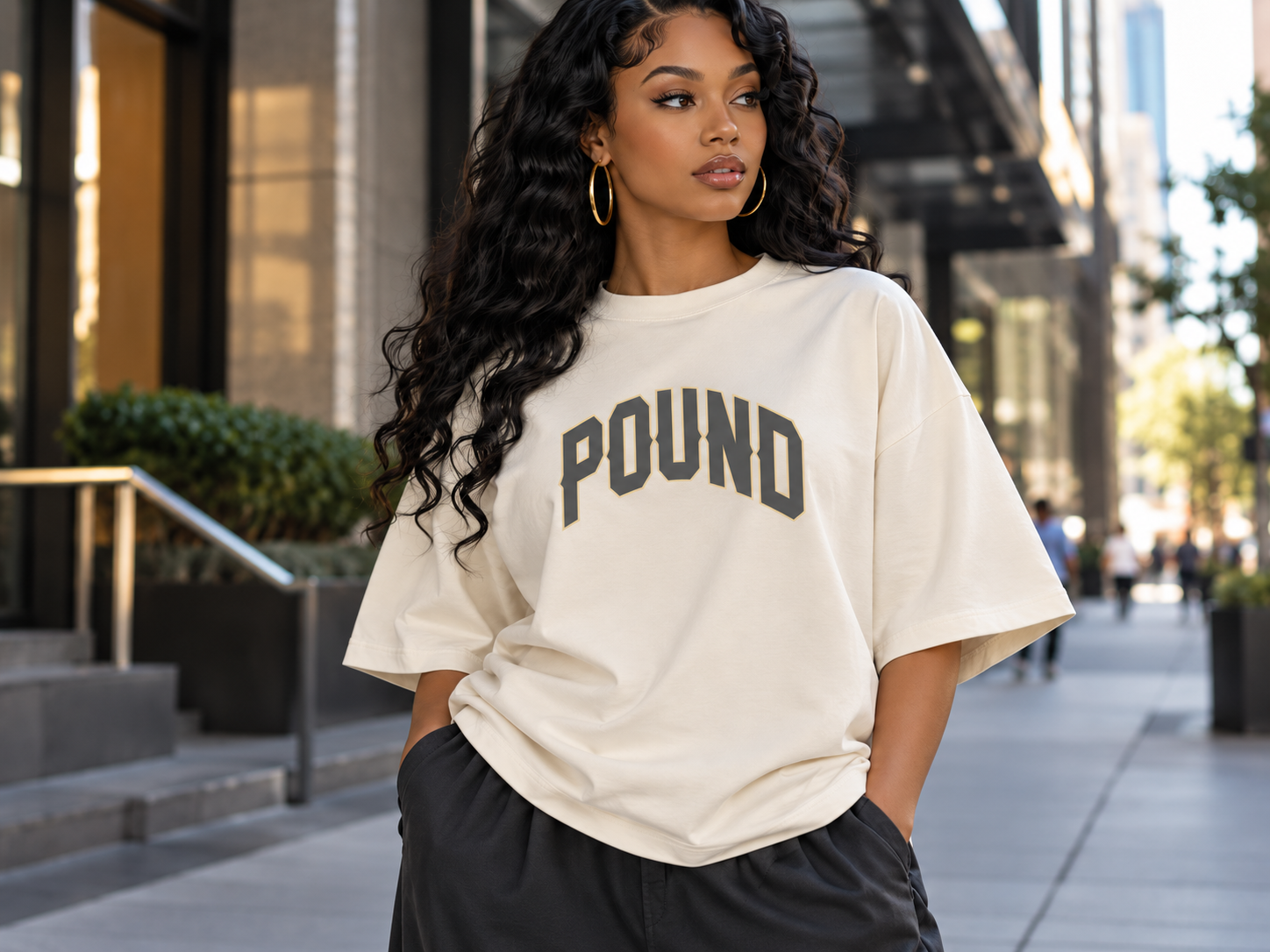 Young woman with long curly black hair wearing a white oversized t-shirt with the word 'POUND' in large black letters across the front, standing outdoors in an urban setting.