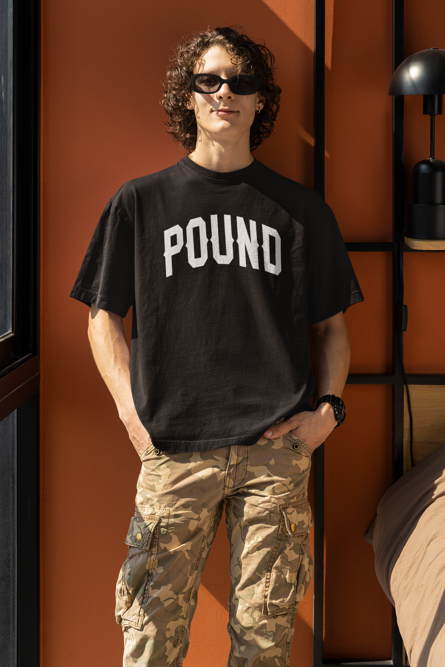 Person with curly hair wearing sunglasses, a black t-shirt reading 'POUND,' and camouflage pants, standing indoors near a wall with a black shelf and lamp.