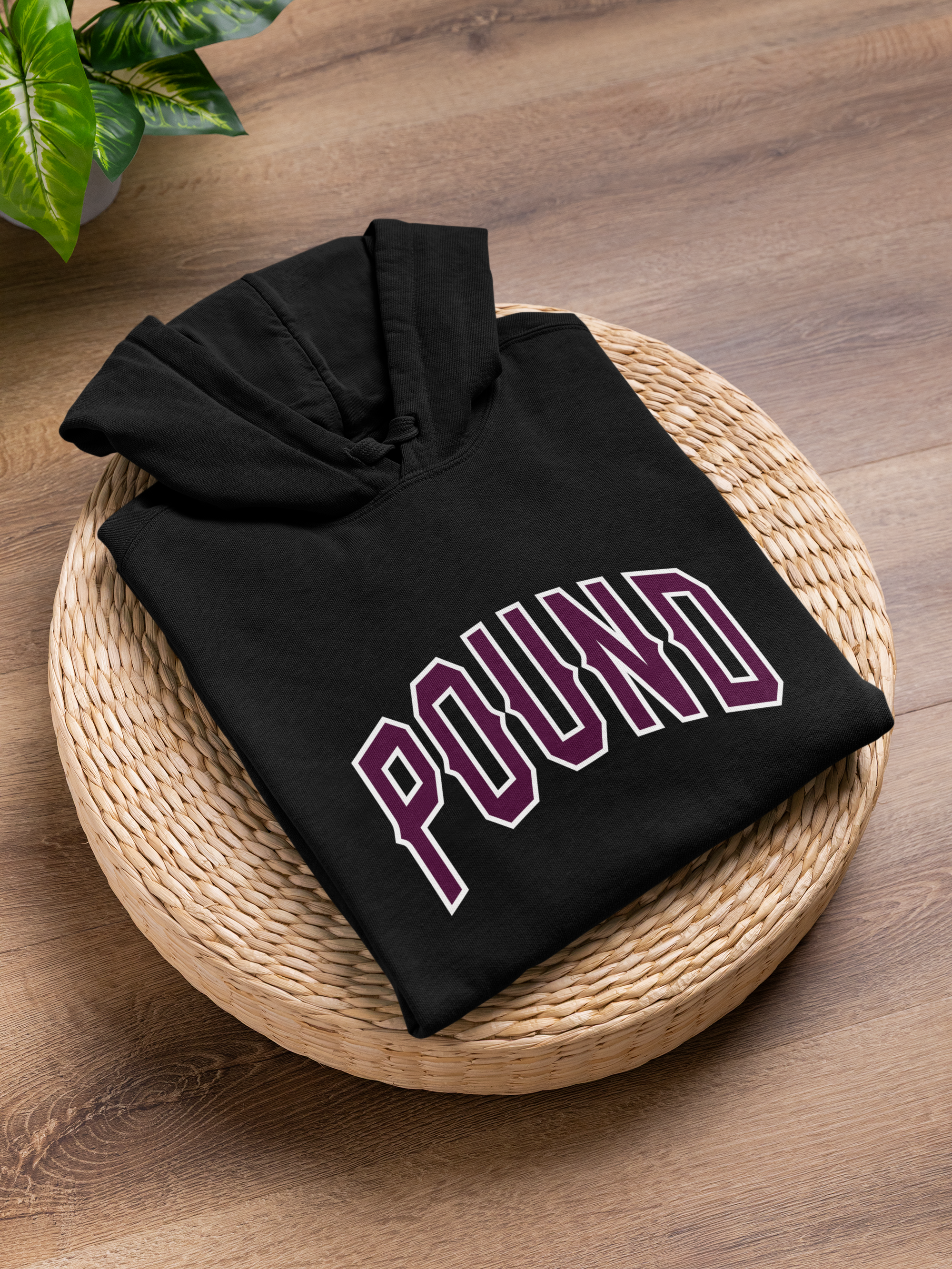 Folded black hoodie with purple and white text, placed on a round woven basket on a wooden surface, with green plant leaves nearby.