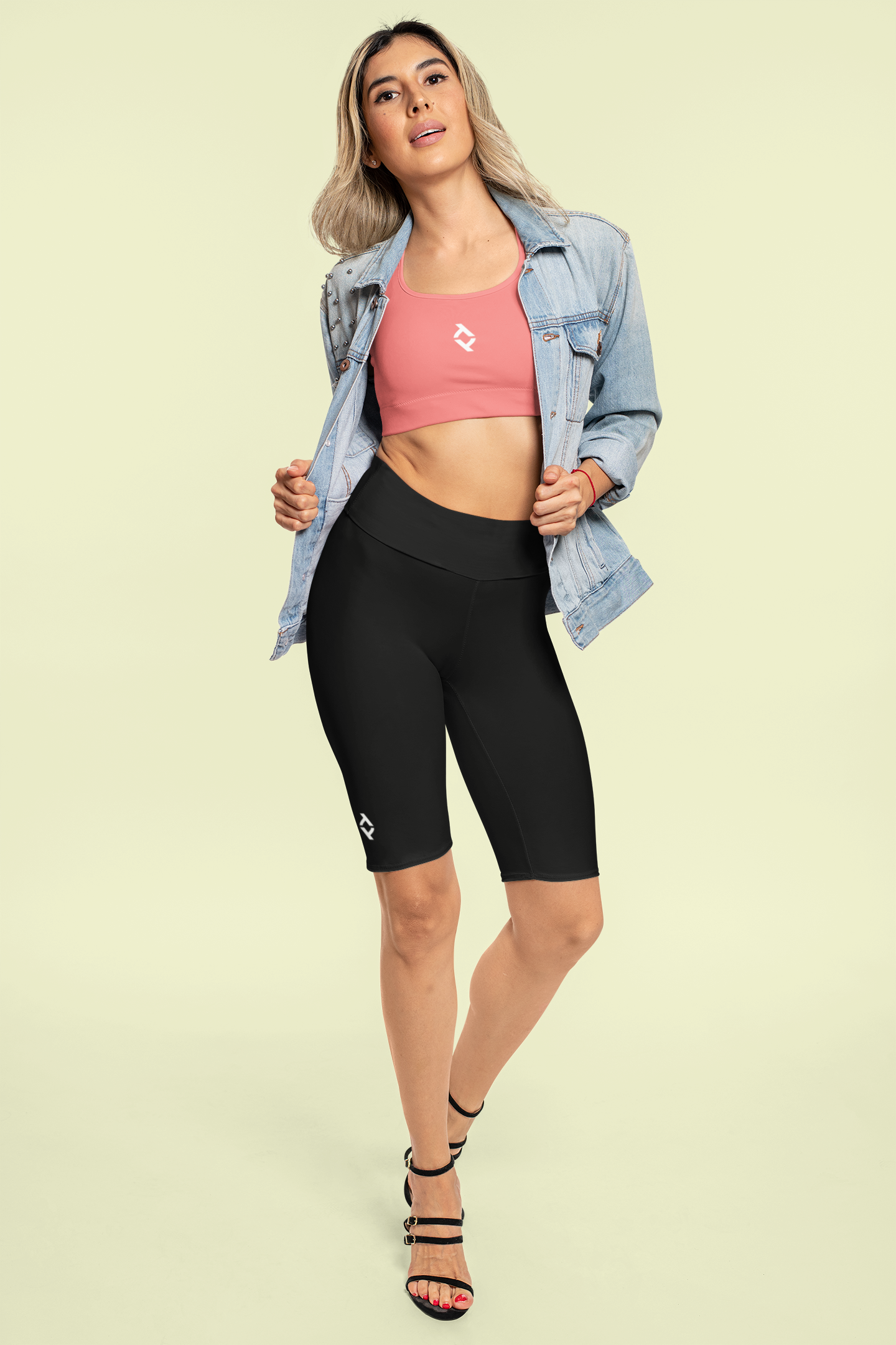 A woman in athletic wear, including a pink sports bra, black athletic shorts, a denim jacket, and black high-heeled sandals, standing against a light green background.
