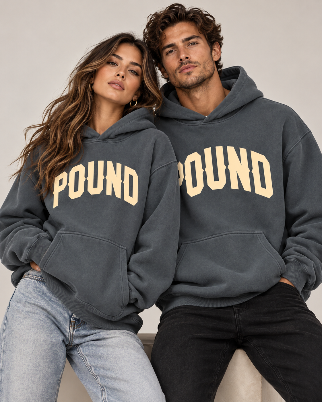 Man and woman wearing matching space gray Pound hoodies with cream varsity logos in a coordinated streetwear studio portrait.