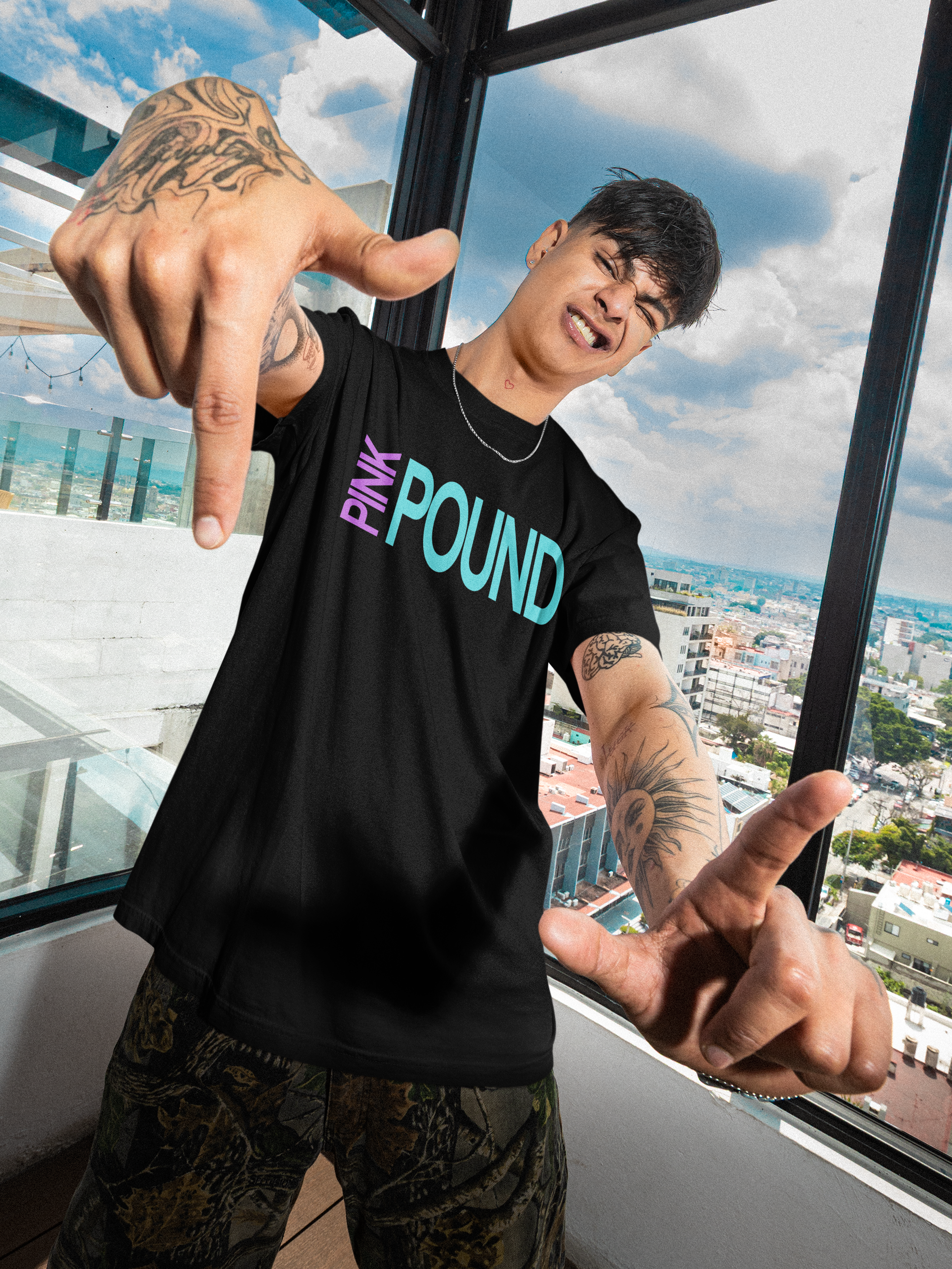 A young man with tattoos on his arms and neck, wearing a black T-shirt with 'Pink POUND' written on it, standing in front of a large window with a cityscape view, making a hand gesture with both hands.