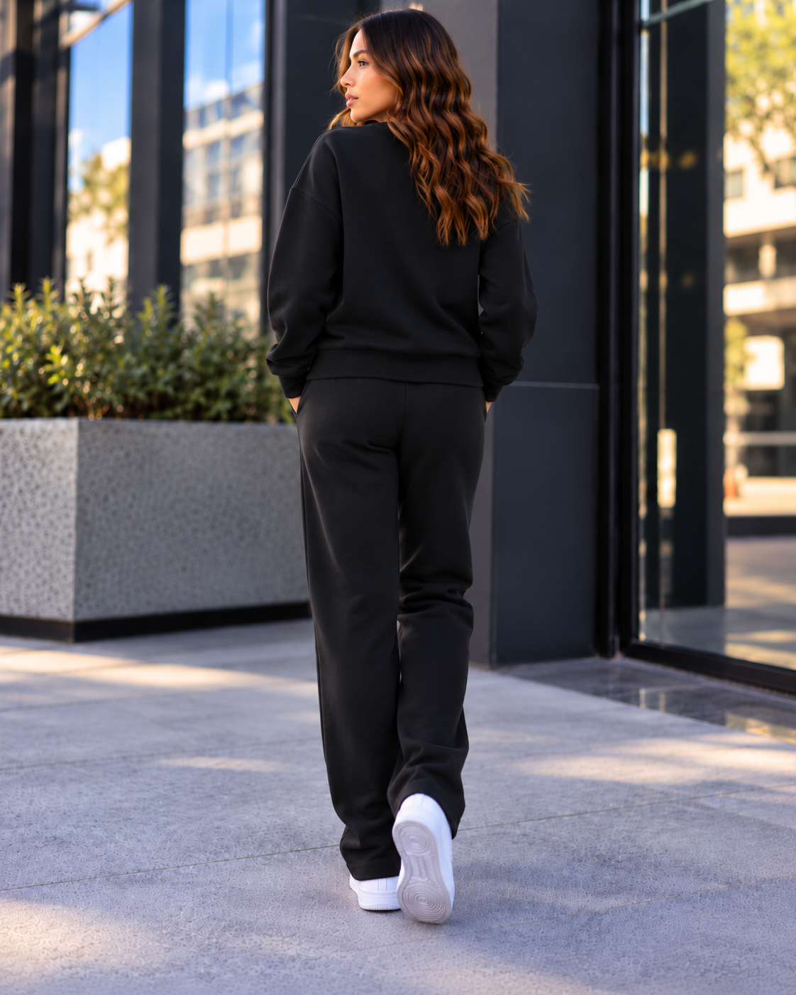 A woman in a black sweatshirt and black pants walking outdoors near modern glass buildings with sunlight reflecting off the windows.