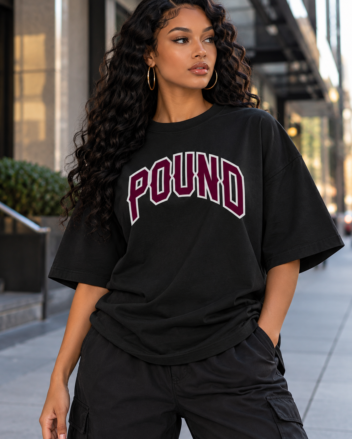 A young woman with long curly hair, gold hoop earrings, wearing a black oversized T-shirt with "POUND" written on it and black pants, stands outdoors in an urban area.