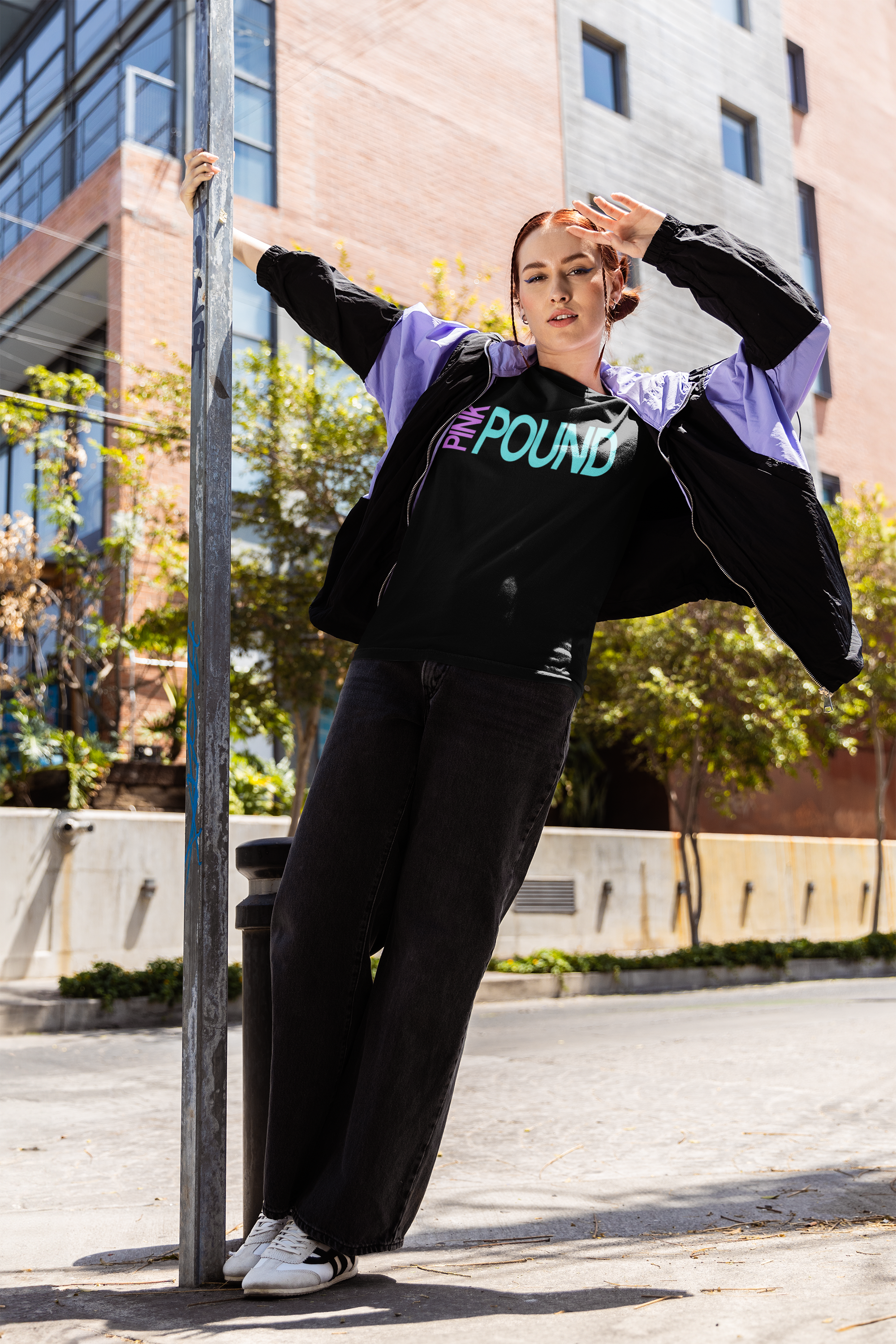 Young woman with red hair in a black t-shirt with colorful text, black wide-leg pants, white sneakers, and a purple and black jacket, standing outside in an urban area with buildings, trees, and sunlight.