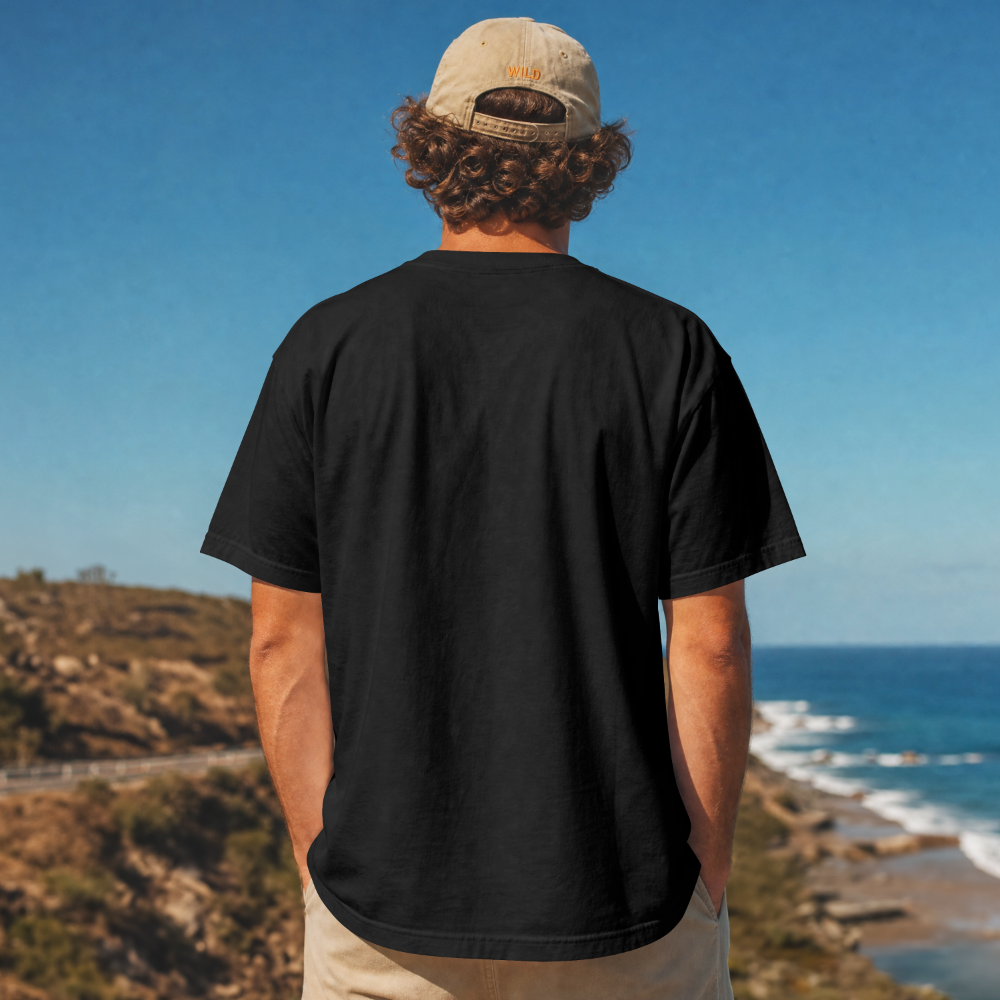 A young man showing back of a premium black t-shirt at the beach with a 100% cotton, 8-ounce fabric weight that is heavy and super soft material. Lightweight feel and breathable for summertime activities.